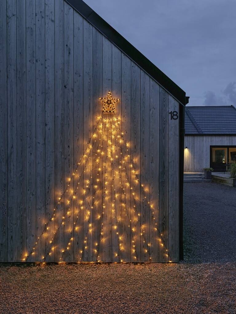 Lichterkette in Form eines Weihnachtsbaums an der Wand eines Hauses angebracht, leuchtet in der Dämmerung auf dunklem Holz.