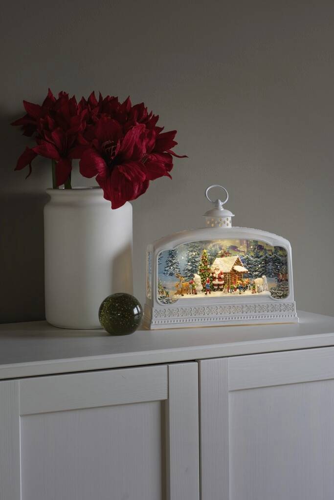 Eine weiße Lampe mit winterlicher Szene zeigt ein Haus im Schnee. Daneben steht eine Vase mit roten Blumen auf einem weißen Schrank.