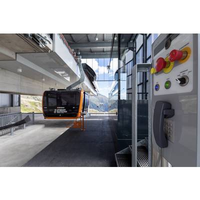 Eine orangefarbene Gondel der Bergbahnstation innen, mit Schalterterminal im Vordergrund. Fenster zeigen eine Berglandschaft draußen.