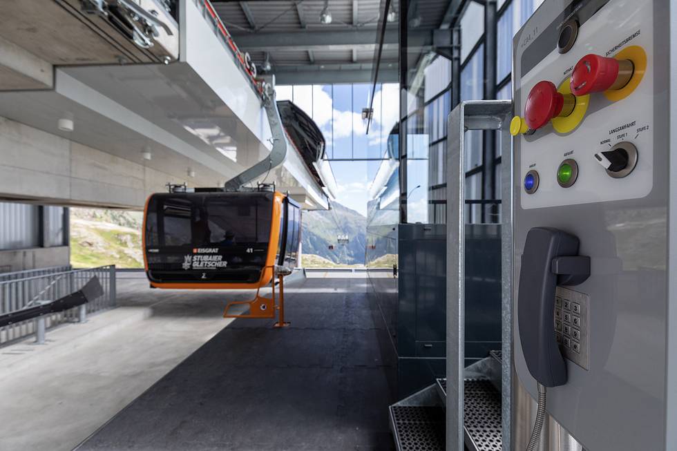 Seilbahnstation Innenansicht mit aufgehängter Gondel, Steuerpult im Vordergrund, Berglandschaft sichtbar durch Fenster im Hintergrund.