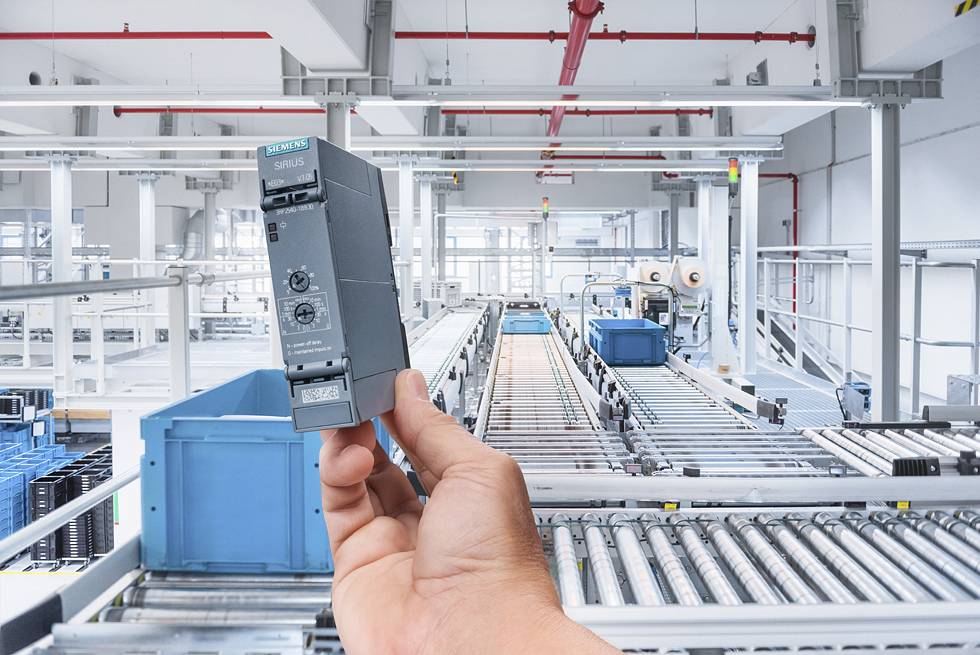 Eine Hand hält ein elektronisches Gerät in einer großen, automatisierten Industrieanlage. Im Hintergrund sind Förderbänder sichtbar.