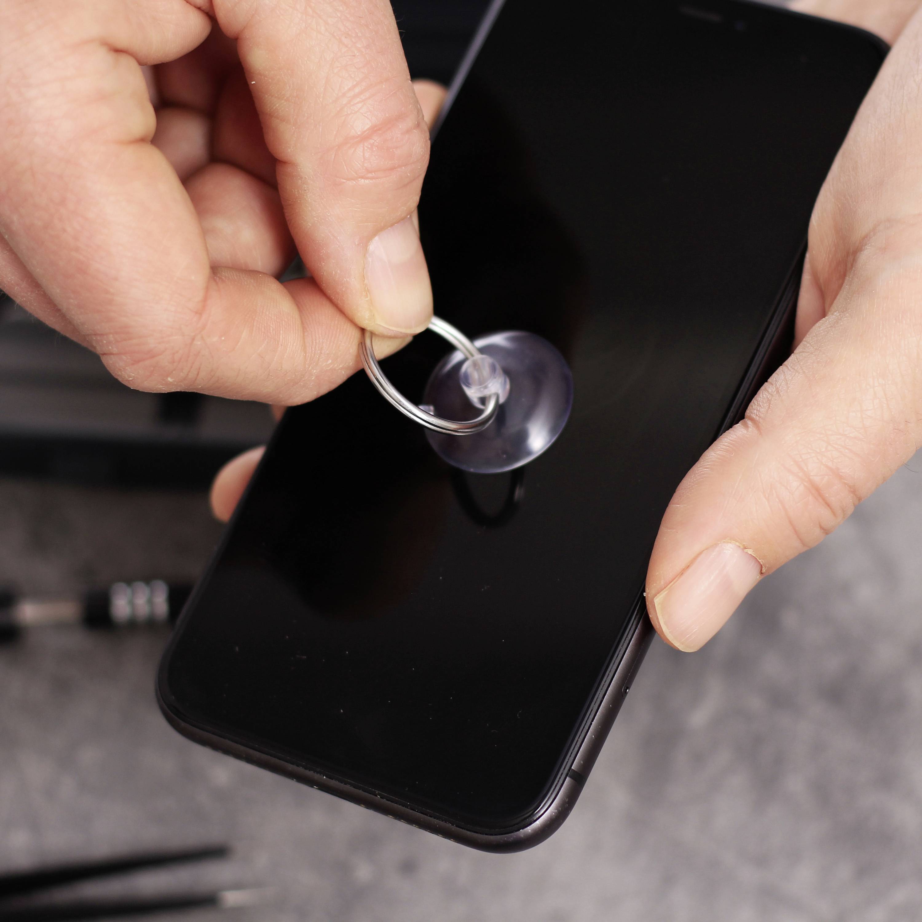Zwei Hände reparieren ein Smartphone mit einem Saugnapf, um das Display abzunehmen. Im Hintergrund liegen Werkzeuge bereit.