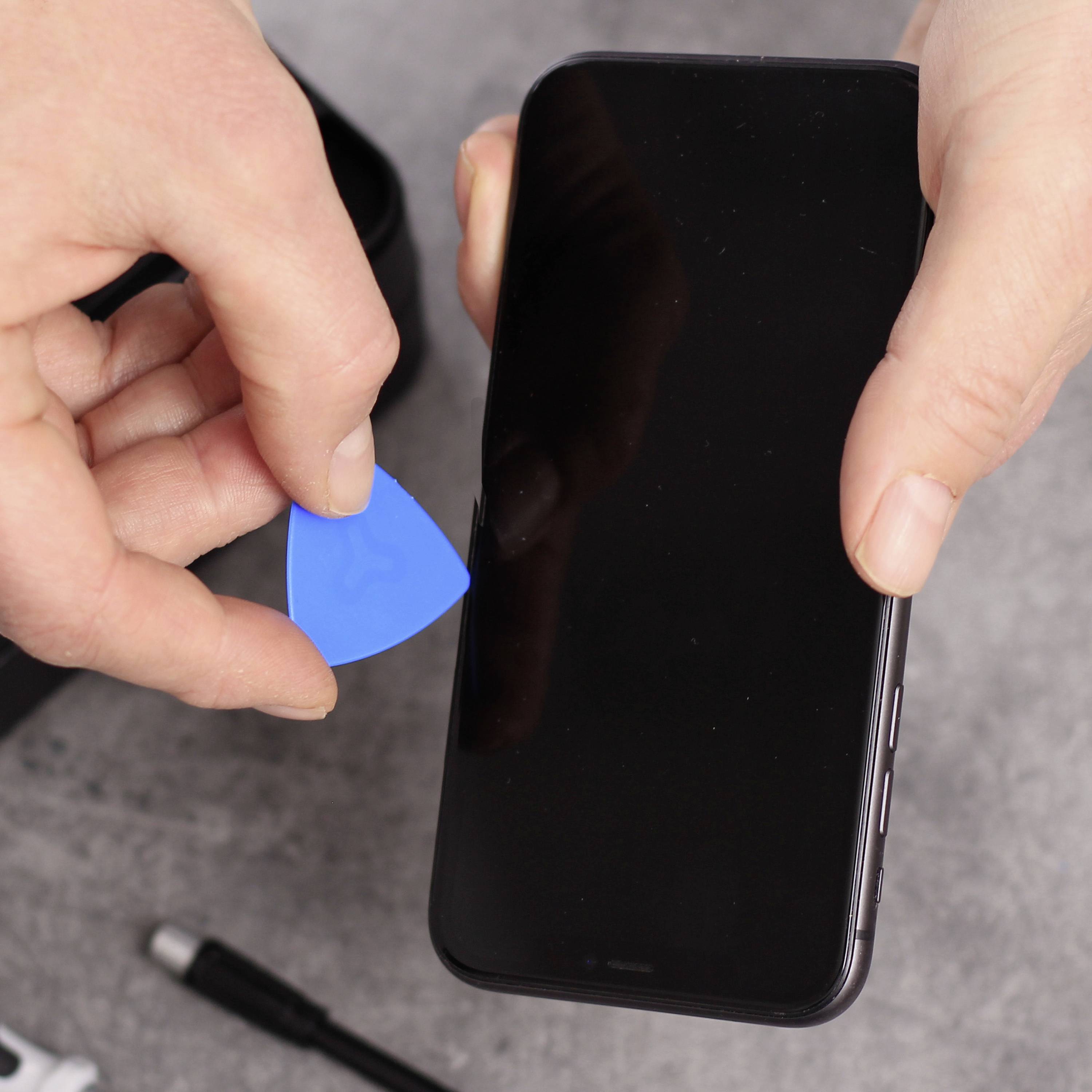 Eine Person hält ein Smartphone und benutzt ein blaues Plastikwerkzeug, um es zu öffnen. Ein Ladekabel liegt auf dem Tisch.