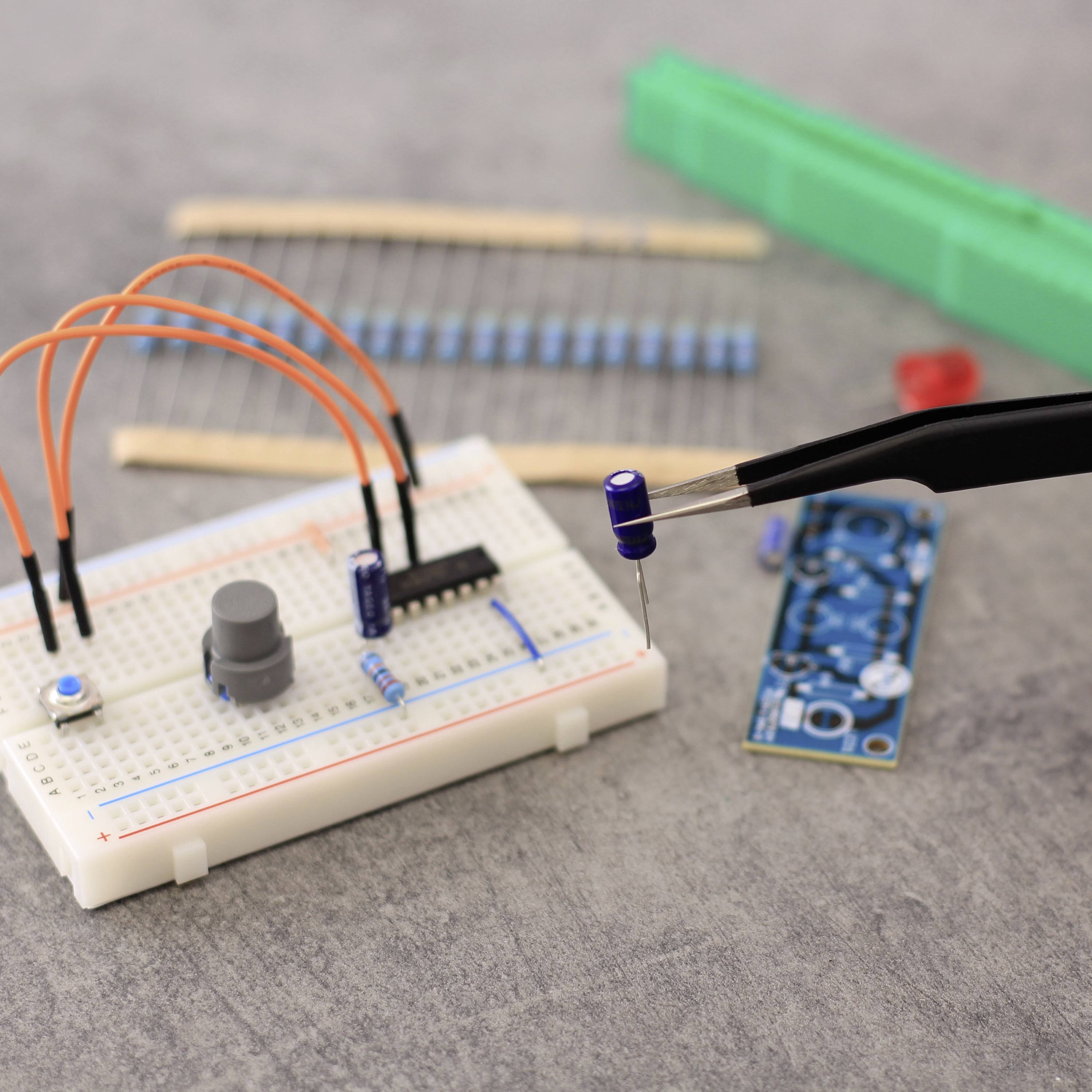 Eine Steckplatine mit Kabeln, Widerständen und einem Mikrocontroller-Chip. Eine Zange hält einen Kondensator. Elektronikbauteile im Hintergrund.