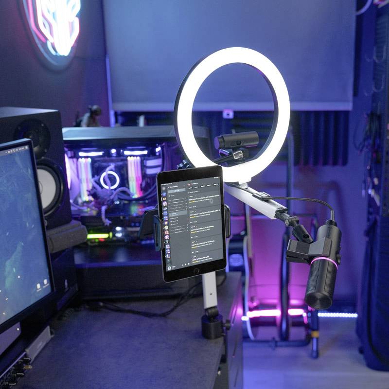 'Streaming-Setup mit Mikrofon, Ringlicht und Tablet auf Schreibtisch; Computer mit leuchtendem Gehäuse im Hintergrund.'