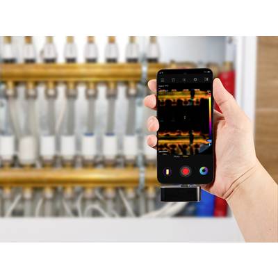 Eine Hand hält ein Smartphone, das ein Wärmebild einer Wasserleitung anzeigt. Im Hintergrund sind unscharfe Rohrleitungen sichtbar.