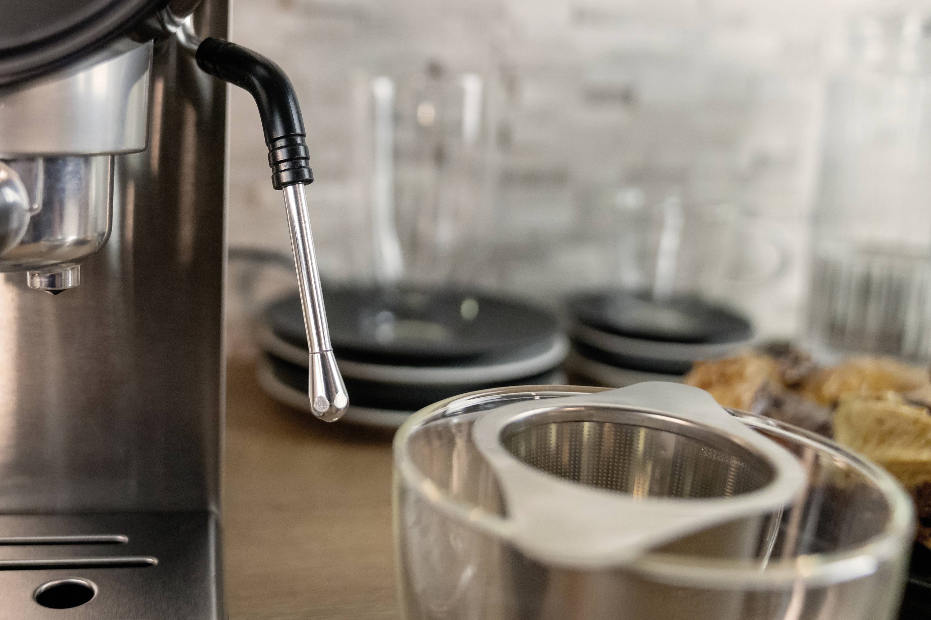 Espressomaschine mit Dampfdüse, dahinter stapelweise Teller, Gläser und ein Teesieb in einer Tasse. Kuchenstücke auf einem Teller.