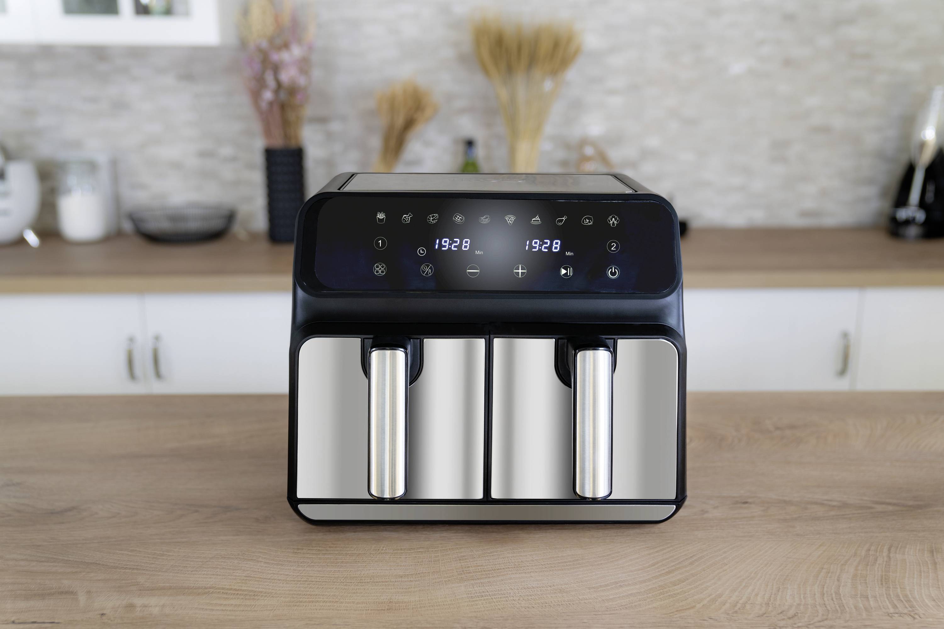 Eine Doppelfritteuse mit digitalem Display, auf einer Küchenarbeitsfläche. Auf dem Display sind Zahlen und Symbole zur Bedienung sichtbar. Im Hintergrund: Küchenutensilien und Dekoration.