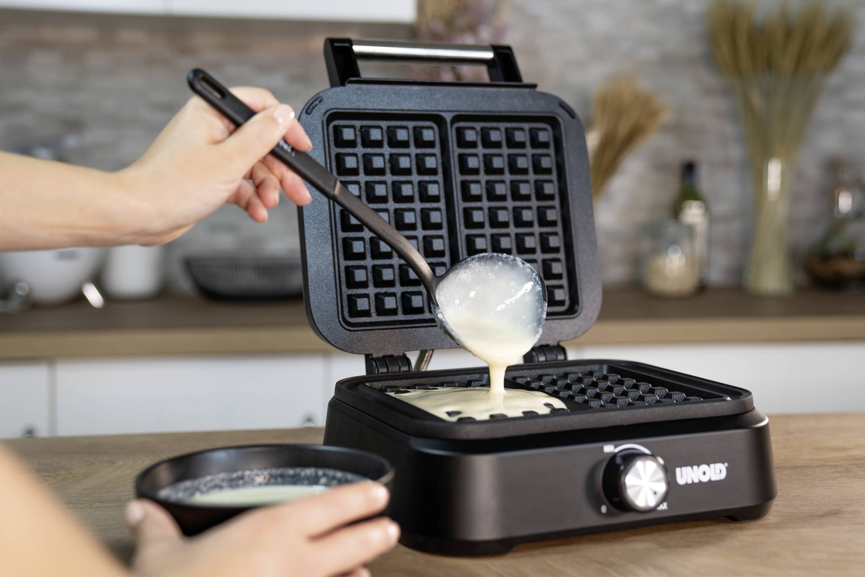 Eine Person gießt Waffelteig mit einer Kelle auf ein offenes Waffeleisen in einer Küche ein. Im Hintergrund sind Küchenutensilien sichtbar.