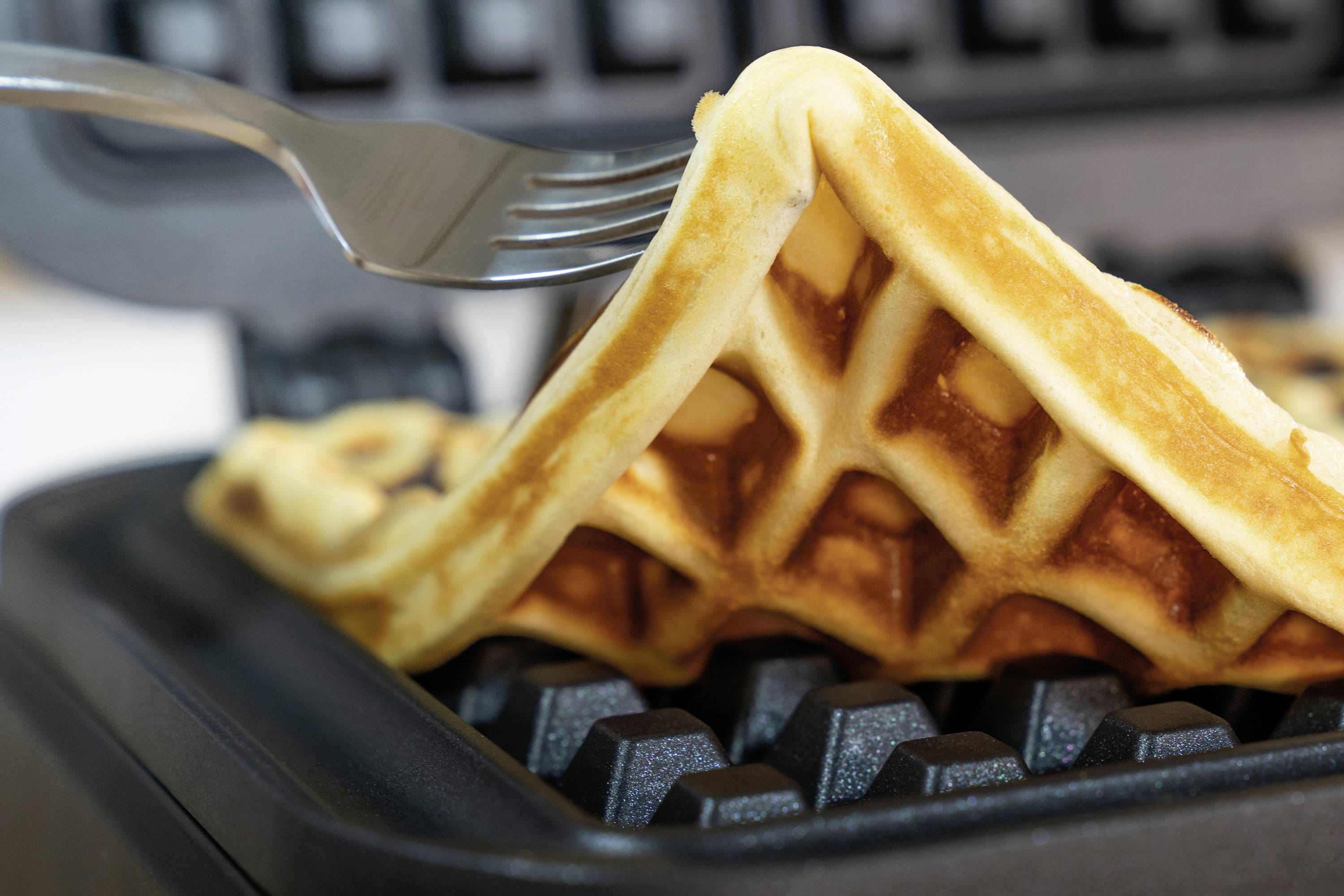 Waffel wird mit einer Gabel aus einem Waffeleisen gehoben. Golden gebräunt und frisch gebacken.
