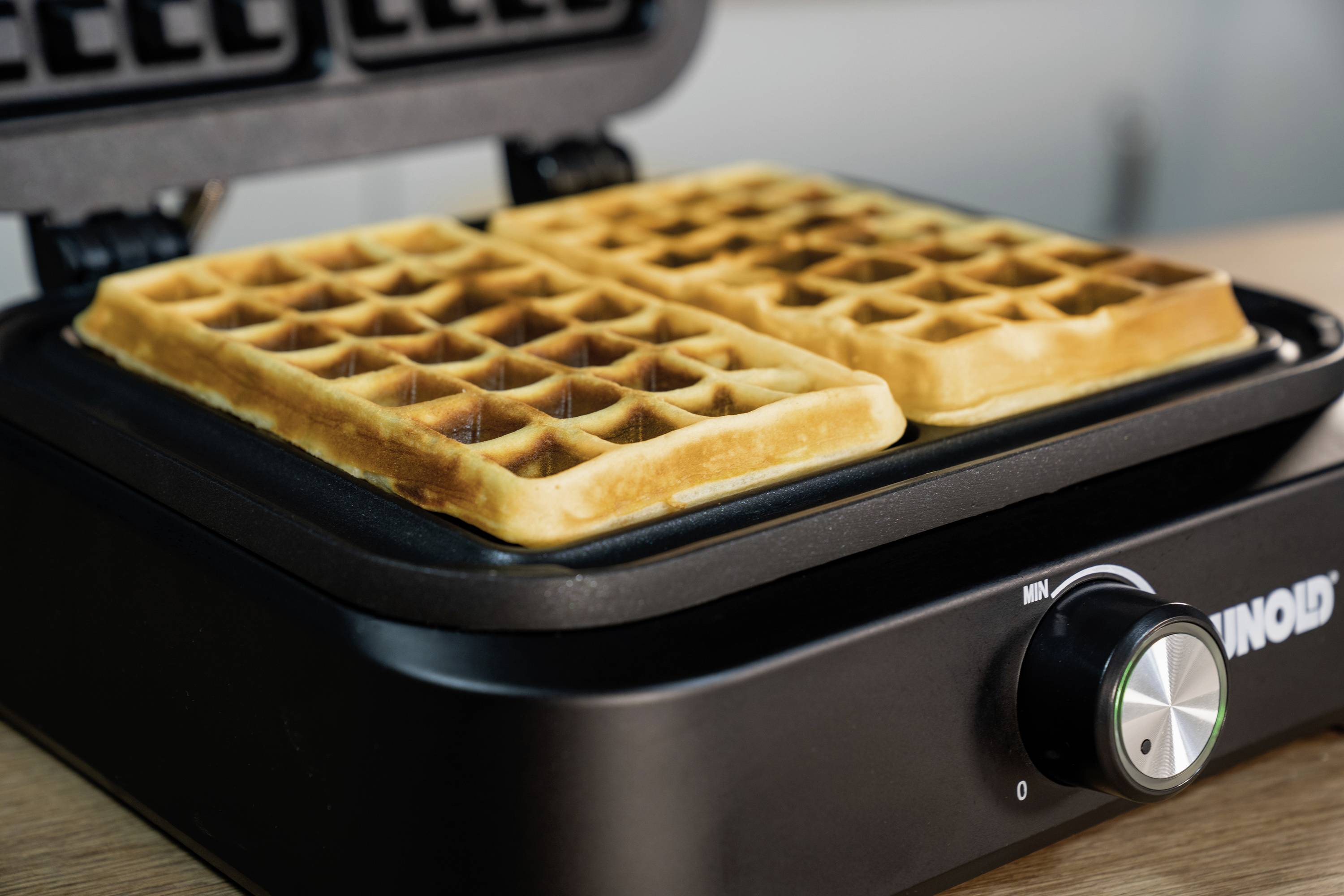Ein Waffeleisen backt zwei goldbraune Waffeln. Der Drehknopf an der Vorderseite des Geräts zeigt die Temperatureinstellung.