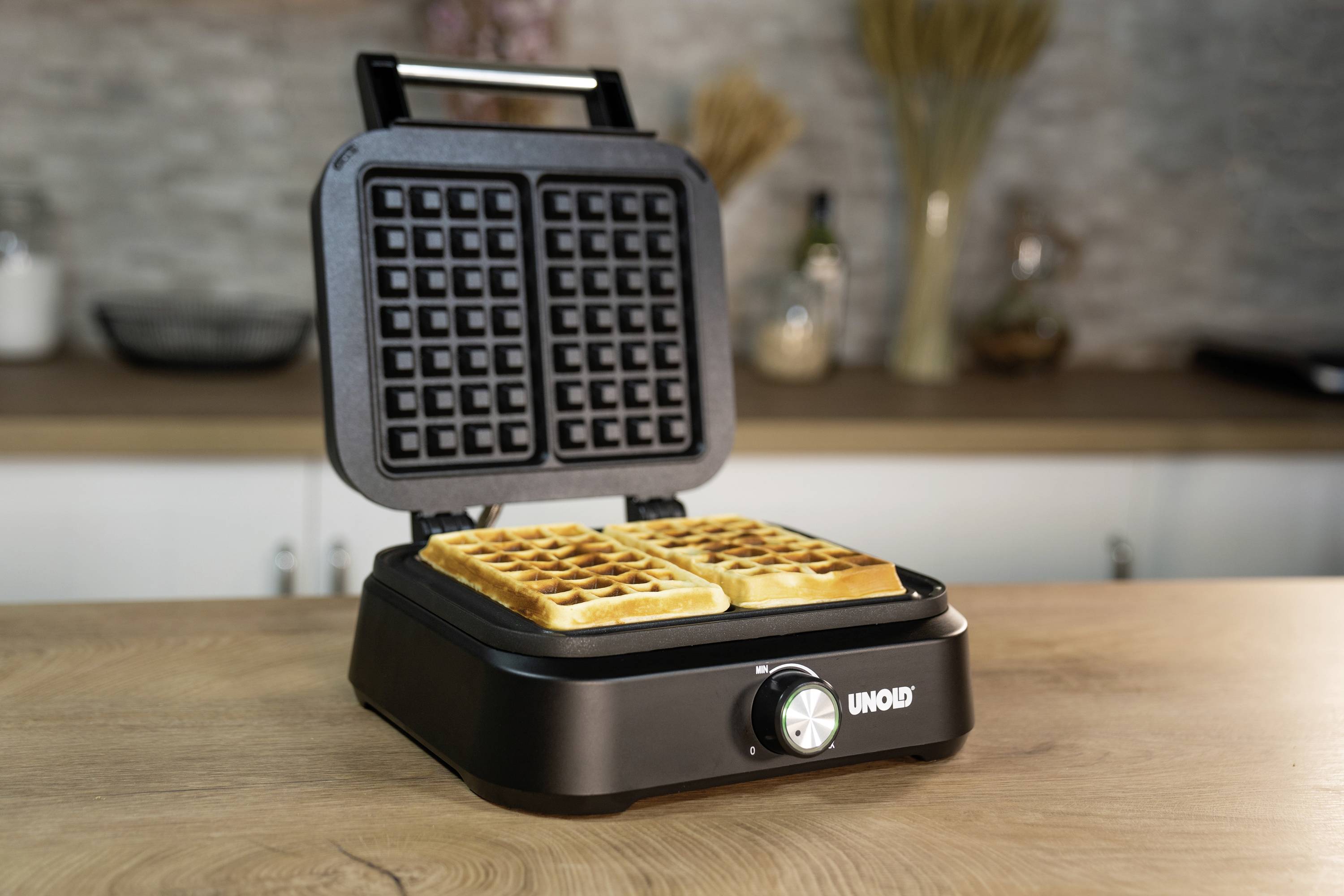Ein offenes Waffeleisen mit zwei gebackenen Waffeln darin, auf einer Küchenarbeitsplatte stehend. Im Hintergrund eine Küche.