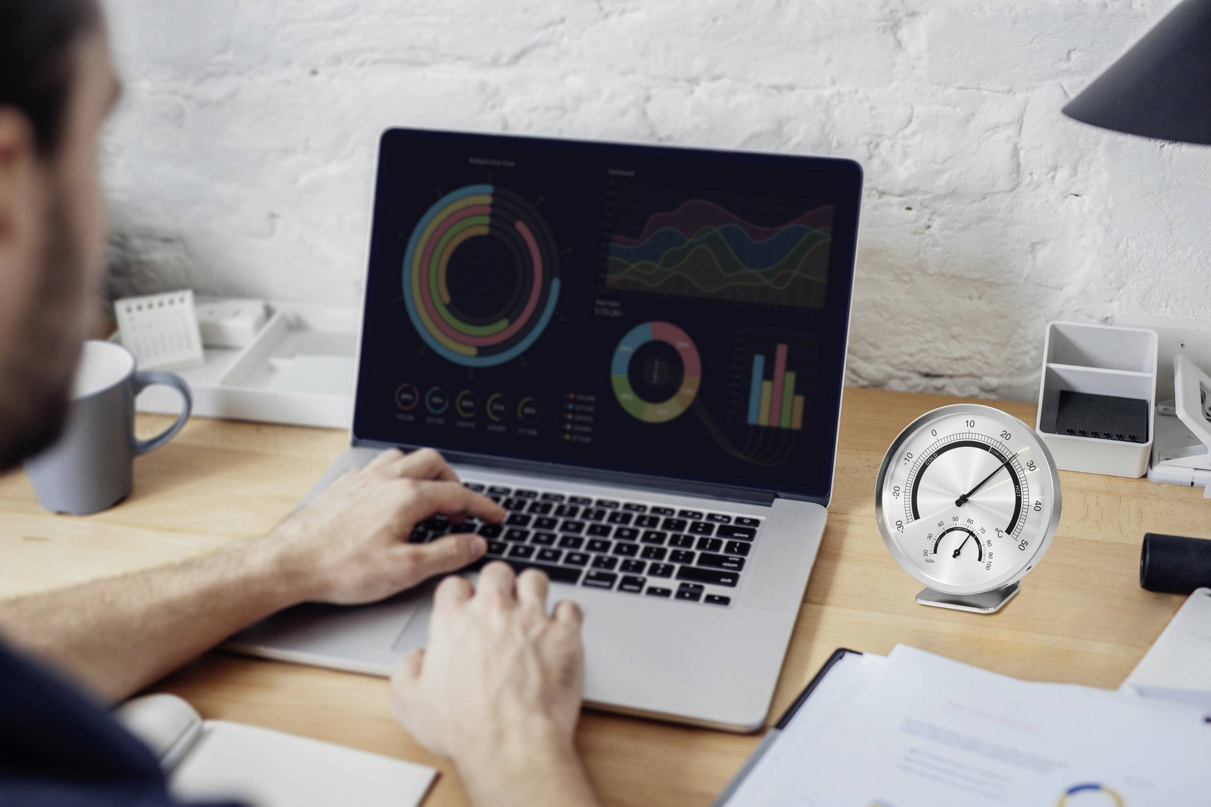 Eine Person arbeitet an einem Laptop mit Datenvisualisierungen und Grafiken. Rechts auf dem Tisch steht ein Tachometer.