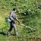 Eine Person in Arbeitskleidung schneidet mit einem Rasentrimmer Gras auf einer grünen Wiese. Im Hintergrund sind Büsche zu sehen.
