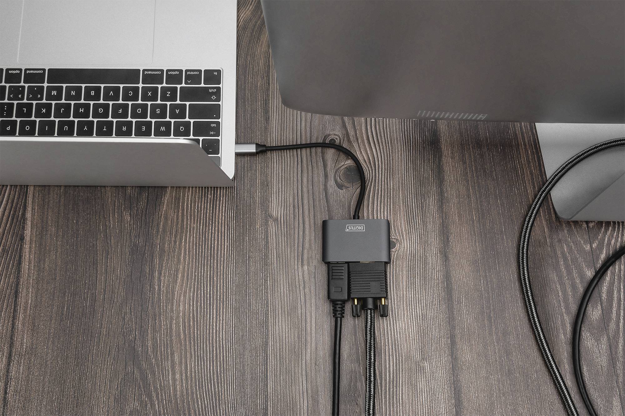 Laptop mit einem Adapter, der über mehrere Kabel mit einem Monitor verbunden ist, auf einer Holzoberfläche.