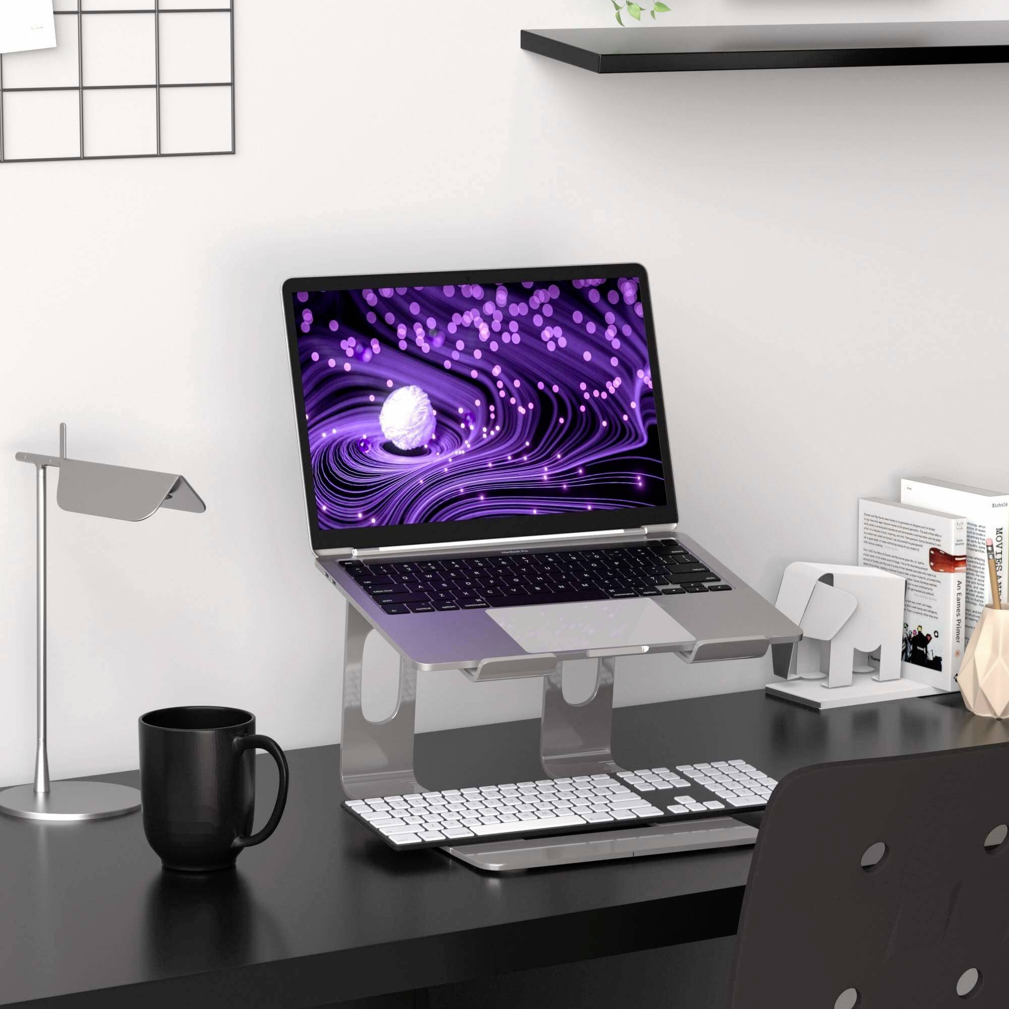 Ein Schreibtisch mit einem Laptop auf einem Ständer. Daneben eine Tasse, Lampe und Bürobedarf. Der Laptop zeigt ein lila Muster.