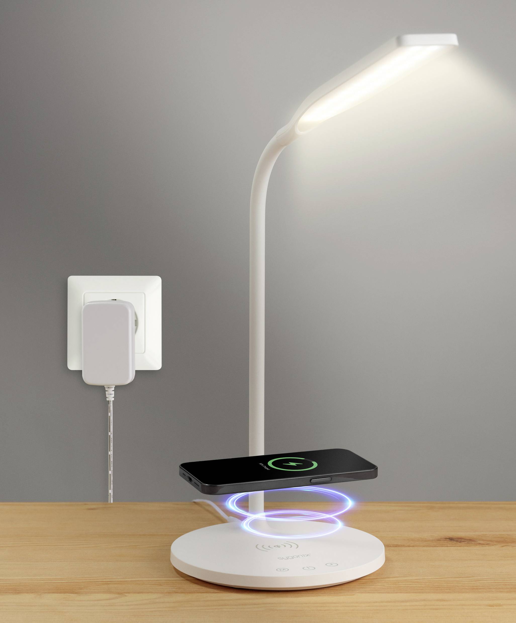 Ein kabelloses Ladegerät auf einem Tisch lädt ein Smartphone unter einer LED-Lampe, die an einer Steckdose angeschlossen ist.