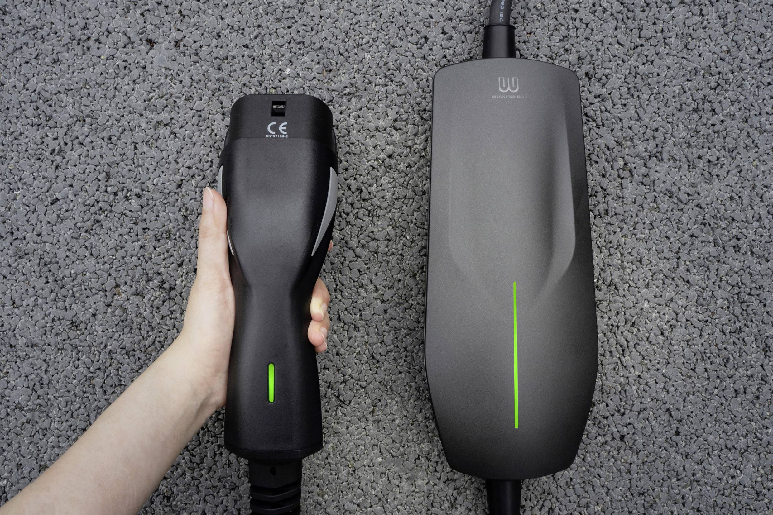 Eine Hand hält den Ladestecker eines Elektroautoladegeräts vor einer grauen Bodenoberfläche. Das Ladegerät zeigt ein grünes Licht.
