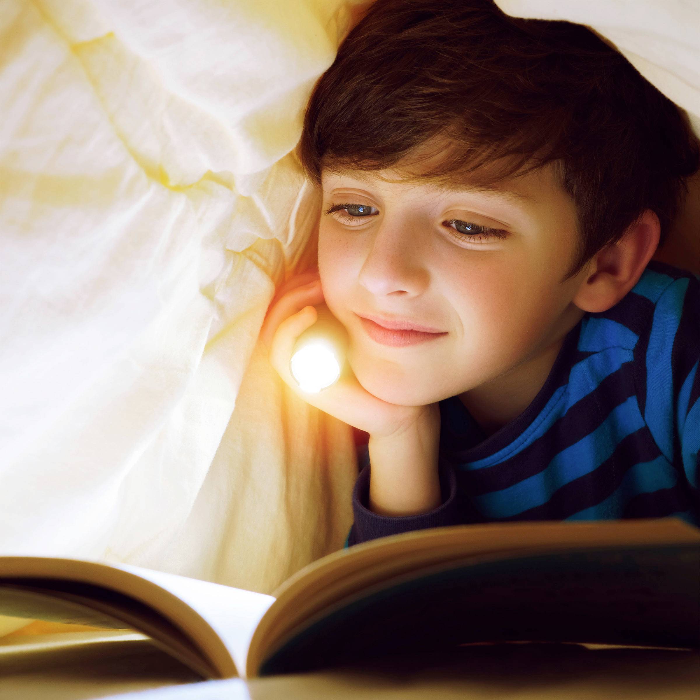 Ein Kind liegt im Bett unter einer Decke und liest ein Buch mit einer Taschenlampe in der Hand und lächelt dabei zufrieden.