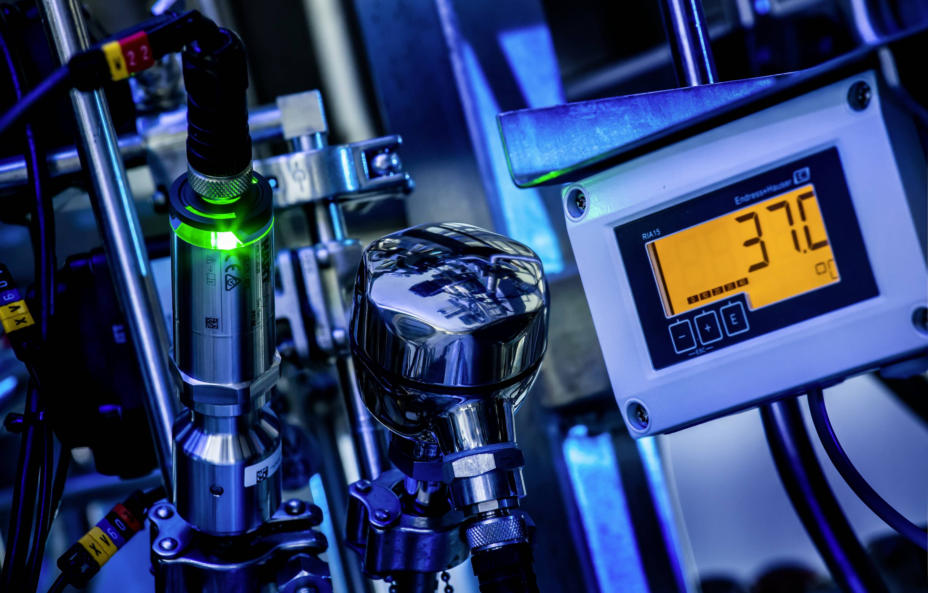 Ein Betriebsdisplay zeigt '37,0°C'. Umgeben von Metallrohren und Anzeigegeräten, deutet die Szene auf eine industrielle Prozessanlage hin.