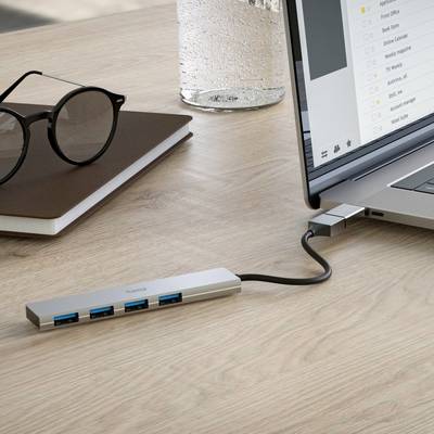 Ein Laptop ist über einen USB-C-Hub mit mehreren USB-Geräten verbunden. Auf dem Tisch liegen eine Brille und ein Notizbuch.