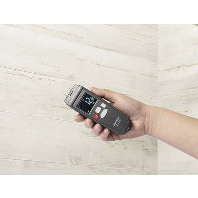 Eine Hand hält ein digitales Feuchtigkeitsmessgerät vor einer hellen, gefliesten Wand. Das Display zeigt '12 %' an.