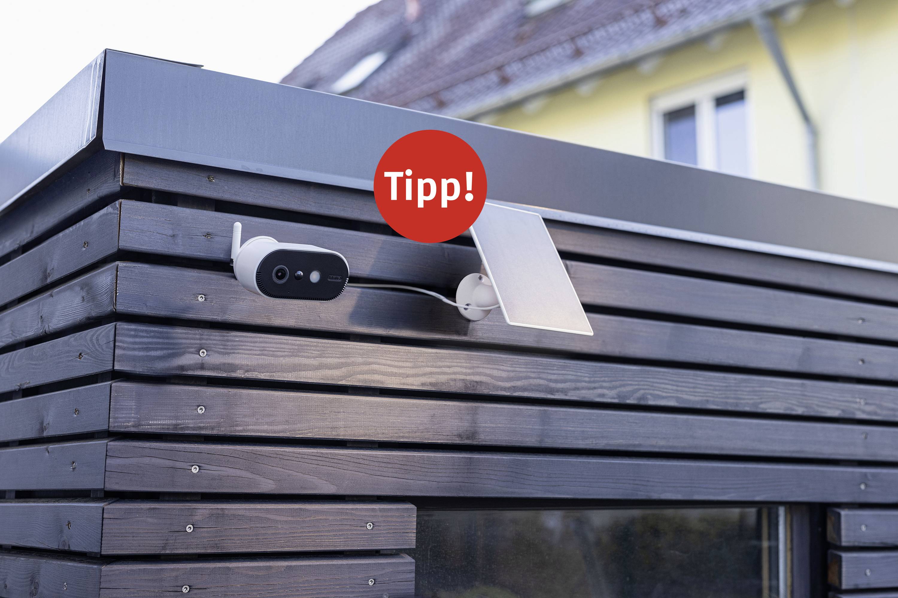Eine Sicherheitskamera ist an einer Holzwand eines Gebäudes montiert. Über der Kamera steht ein roter Kreis mit 'Tipp!'.