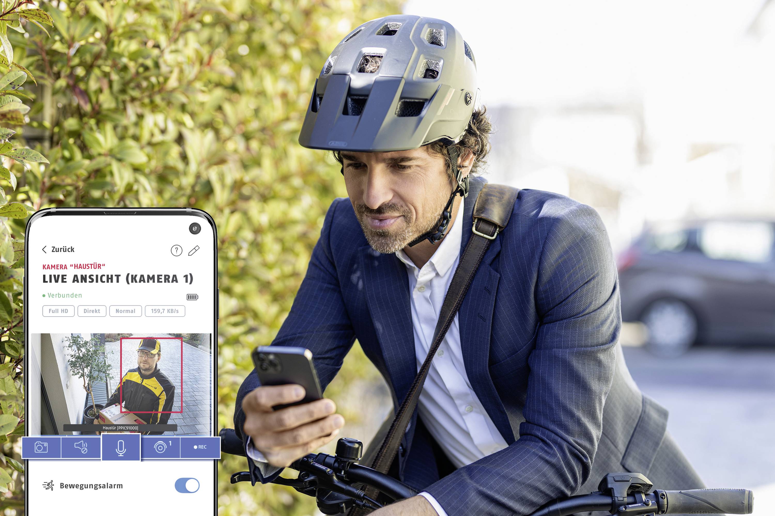Mann in Anzug und Helm schaut auf sein Smartphone neben Fahrrad. Auf dem Handybildschirm wird eine Live-Übertragung einer Überwachungskamera angezeigt.