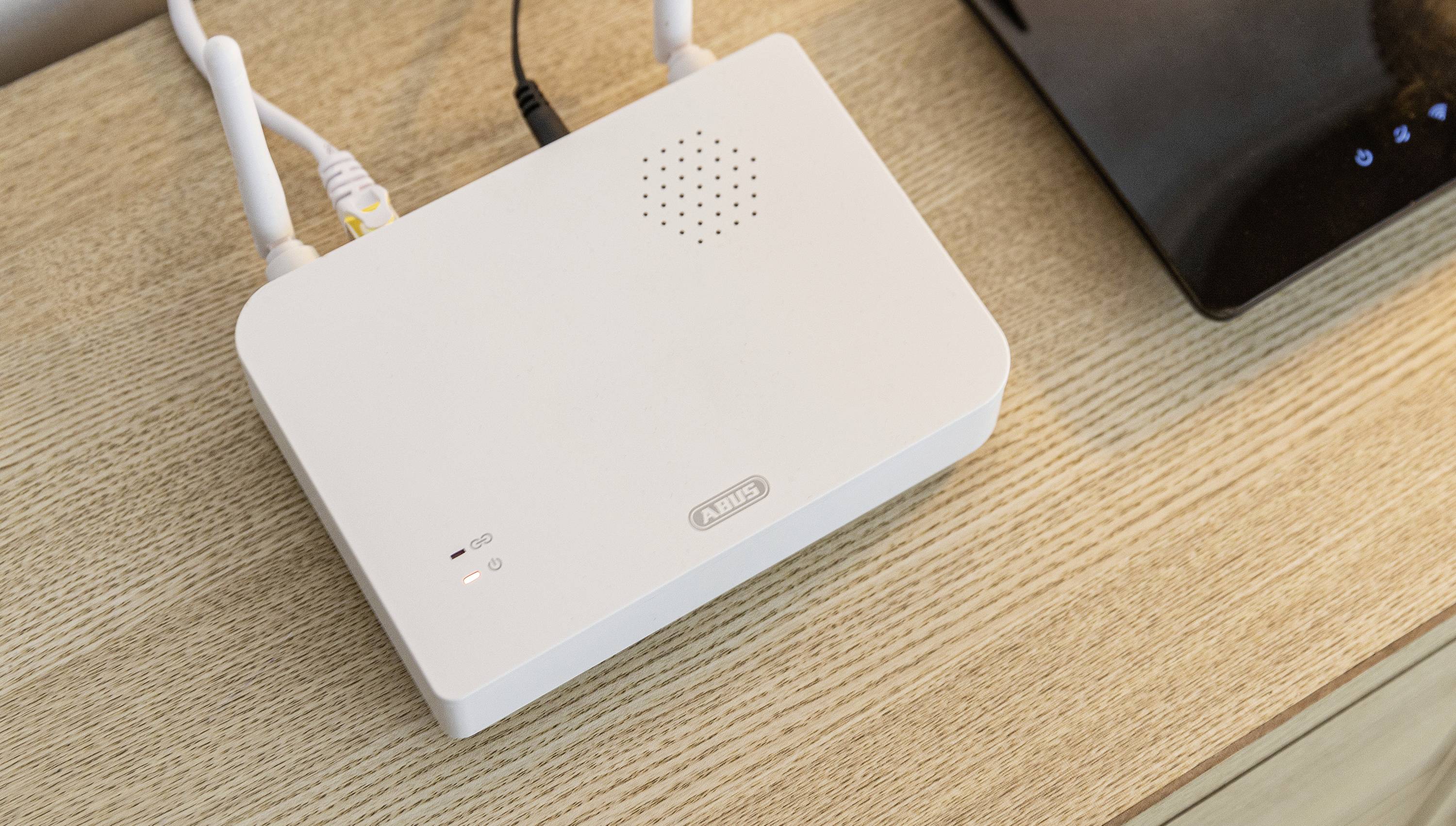 Ein weißer WLAN-Router mit zwei Antennen steht auf einem Holztisch. Daneben befindet sich ein schwarzes Gerät.