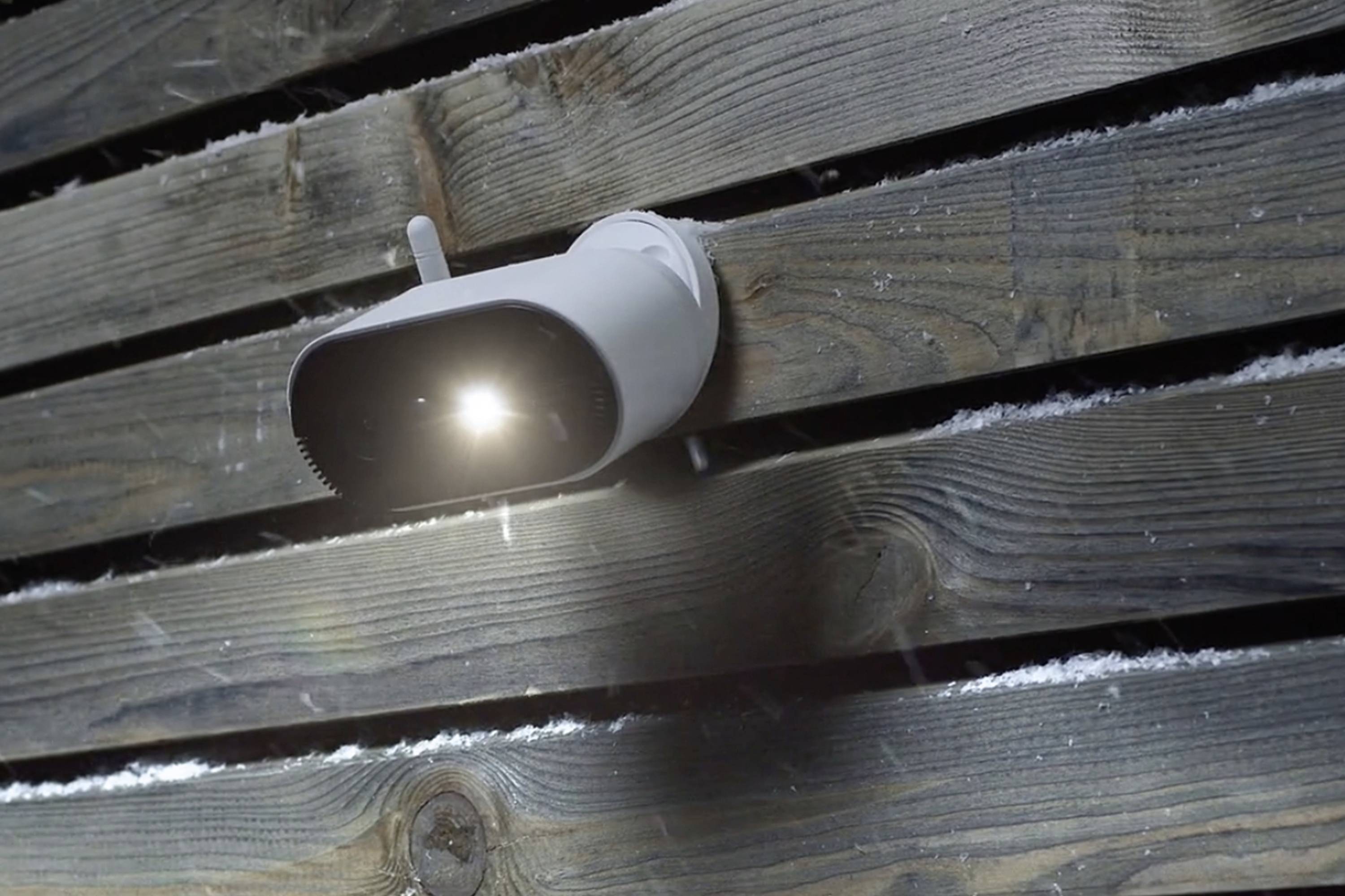 Eine weiße Sicherheitskamera mit eingeschaltetem Licht ist an einer dunkel gebeizten Holzfassade montiert. Einige Schneeflocken sind sichtbar.