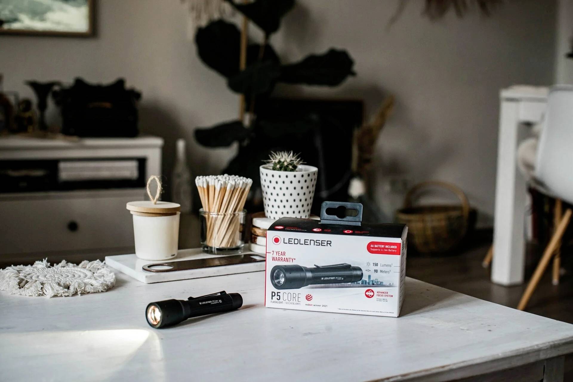 Taschenlampe 'Ledlenser P5 Core' und Verpackung auf weißem Tisch in Wohnzimmer. Im Hintergrund stehen Pflanzen und Dekorationen.
