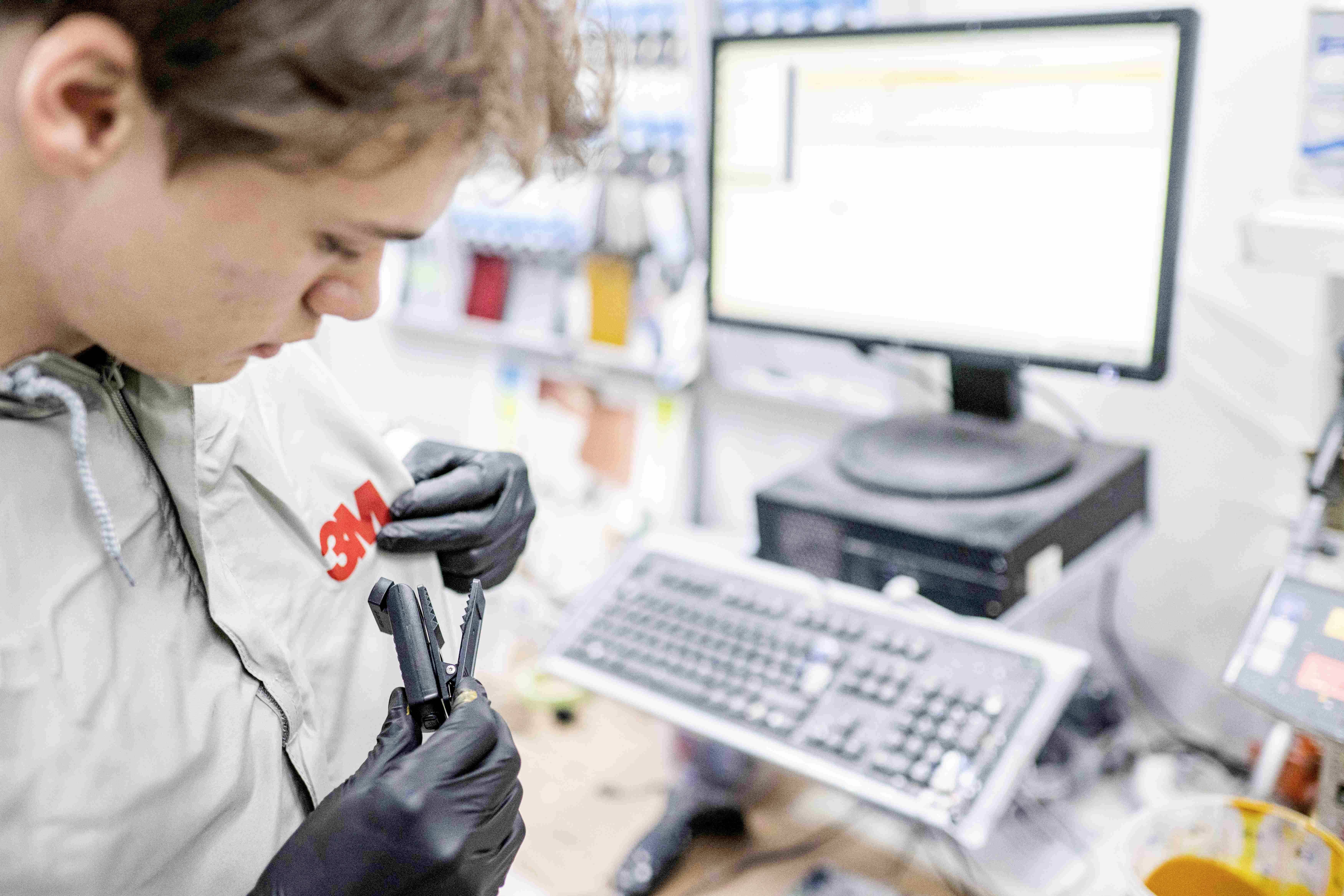 Eine Person in Werkstattkleidung arbeitet konzentriert mit Werkzeugen vor einem Computerbildschirm und einer Tastatur.