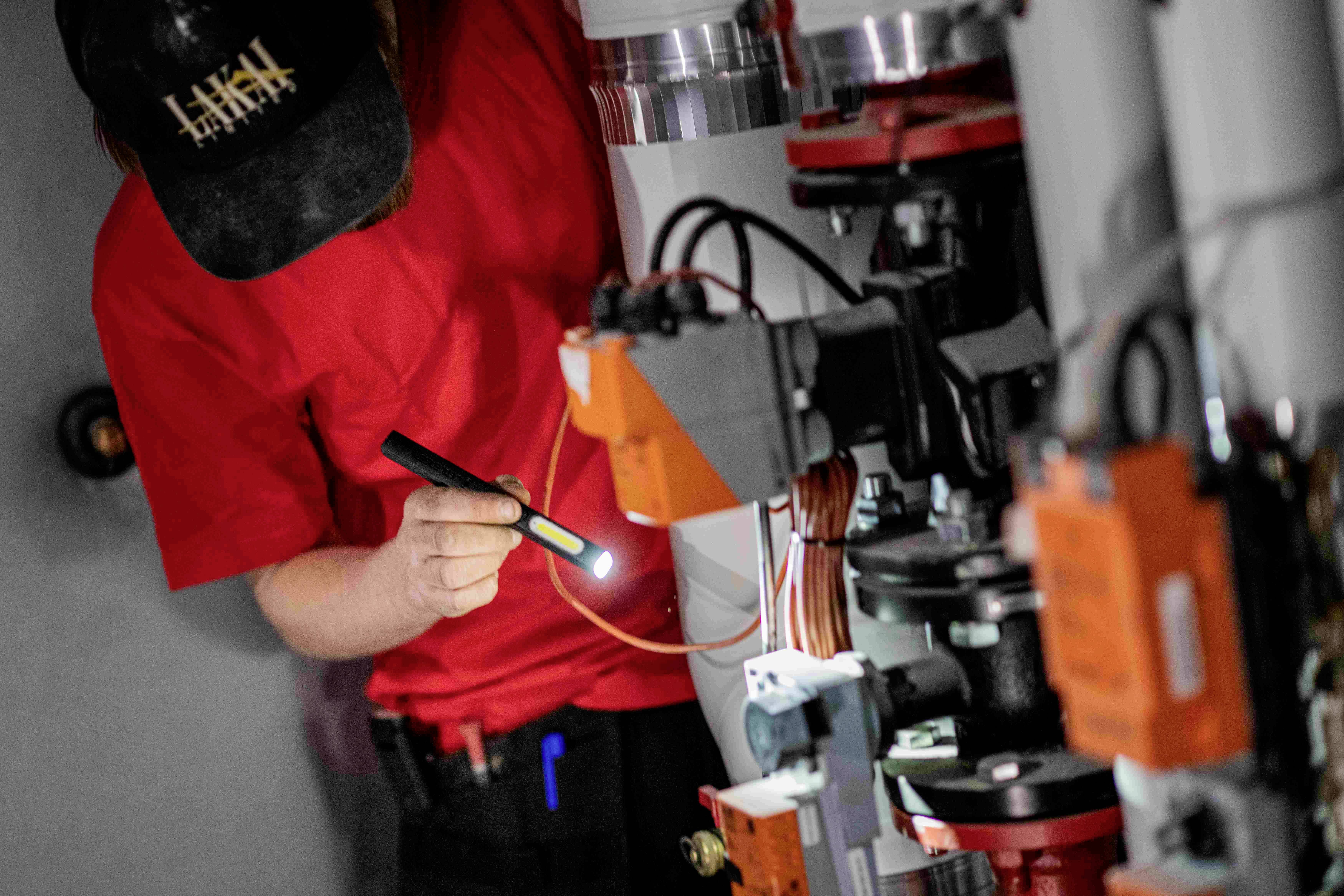 Eine Person in rotem Shirt prüft technische Geräte mit einer Taschenlampe. Elektrische Kabel und Mechanismen sind sichtbar.