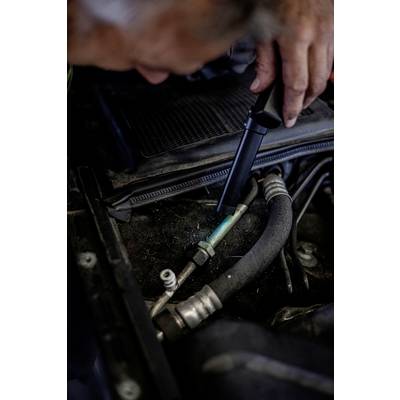 Ein Mechaniker inspiziert einen Autoteil im Motorraum mit einer Taschenlampe. Fokus auf Schläuche und Verbindungen.