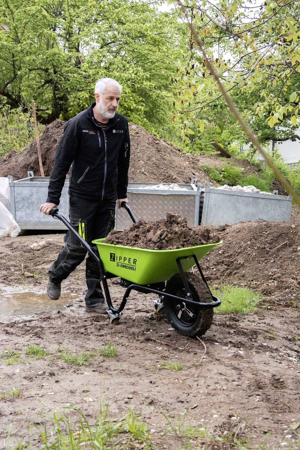 Eine Person schiebt eine grüne Schubkarre voller Erde über einen matschigen Boden in einem Garten oder Baustelle, umgeben von Bäumen und Erdehaufen.