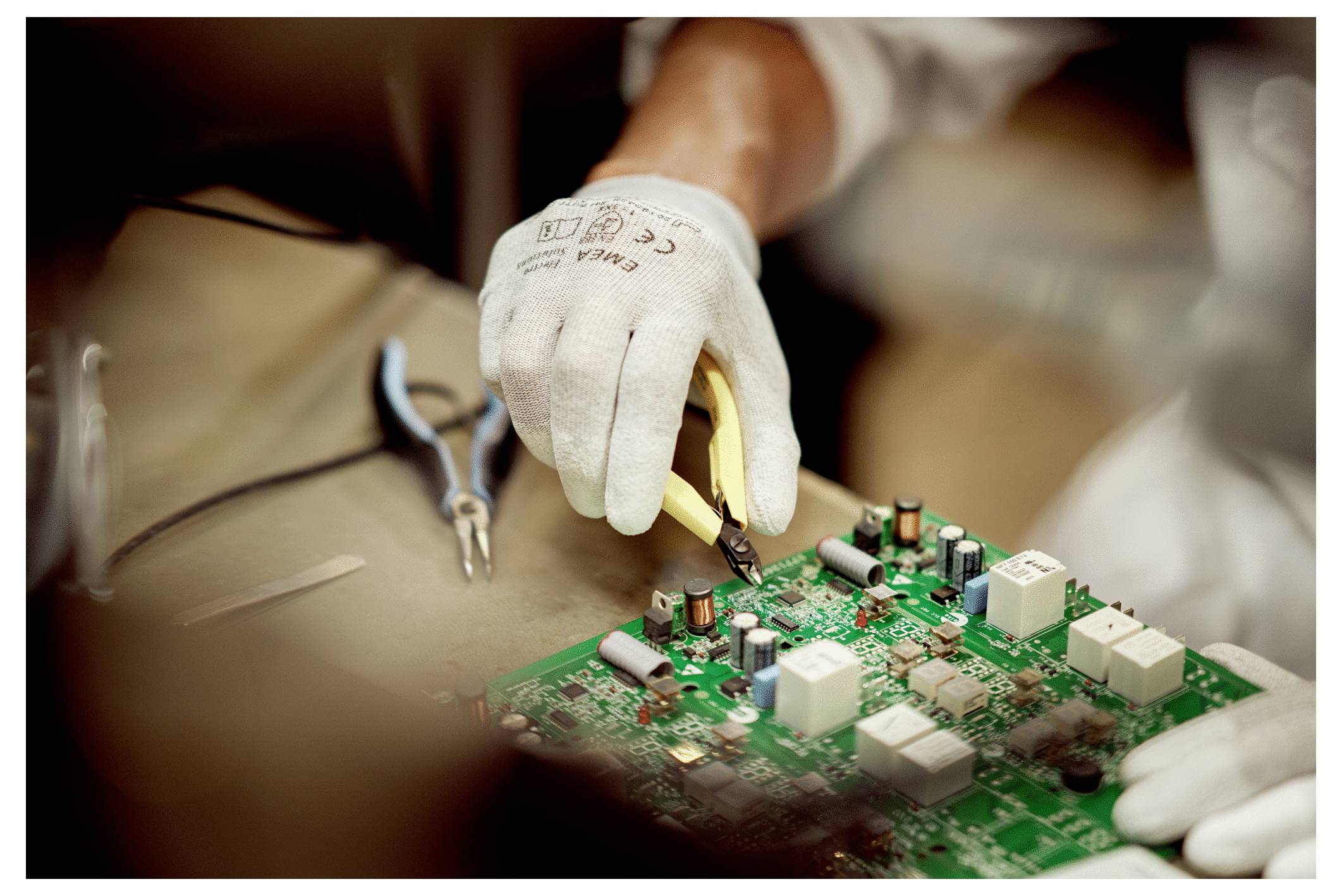 Eine Person mit Handschuhen verwendet einen Lötkolben auf einer grünen Leiterplatte, wobei Werkzeuge wie Zangen in der Nähe liegen und eine Konzentration auf elektronische Arbeiten andeuten.