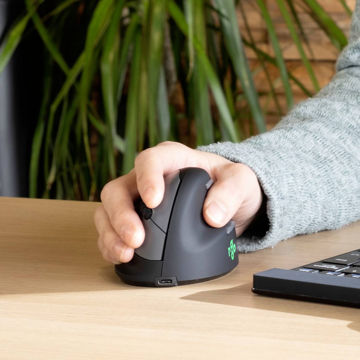Hand hält eine vertikale ergonomische Maus auf einem Tisch neben einer Tastatur. Im Hintergrund sind Pflanzen zu sehen.