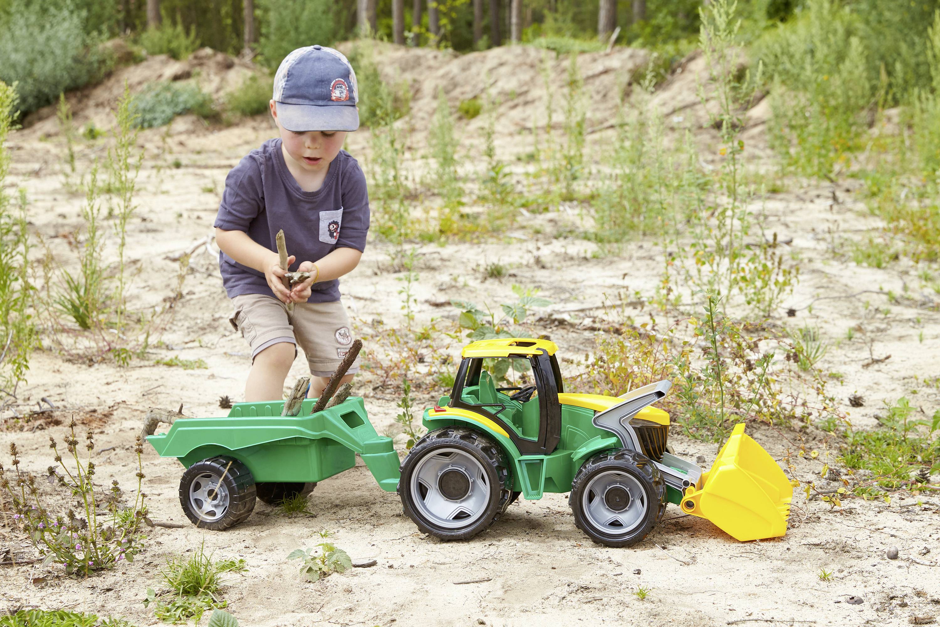 Ein Kind spielt auf sandigem Boden mit einem grünen Spielzeugtraktor und Anhänger. Im Hintergrund sind Gras und Bäume zu sehen.