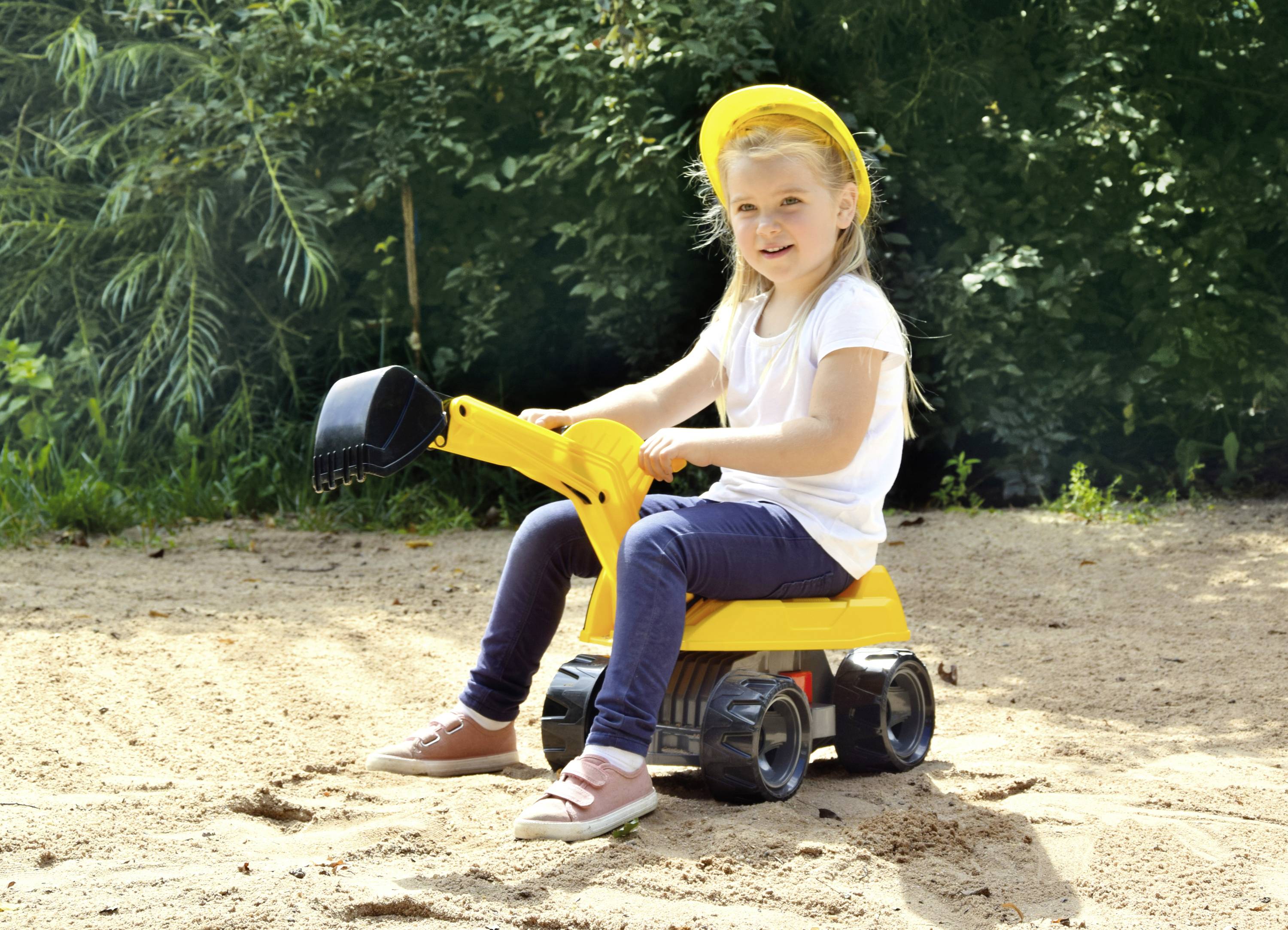 Ein Kind in weißem T-Shirt und blauer Hose sitzt auf einem gelben Spielzeugbagger im Sand und trägt einen gelben Bauhelm.