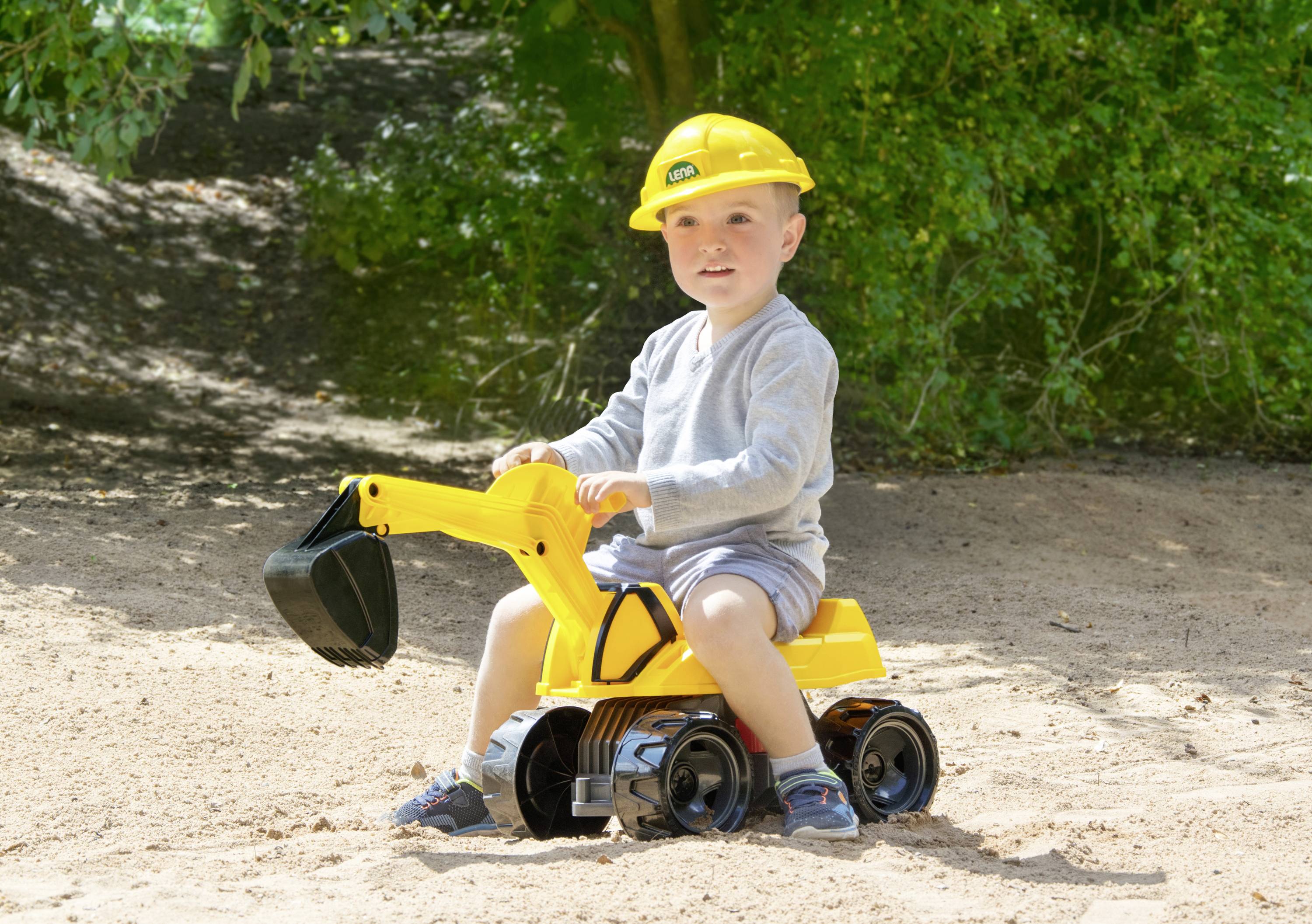 Ein Kind mit Bauhelm sitzt auf einem gelben Spielzeugbagger im Sand, inmitten einer grünen Umgebung.