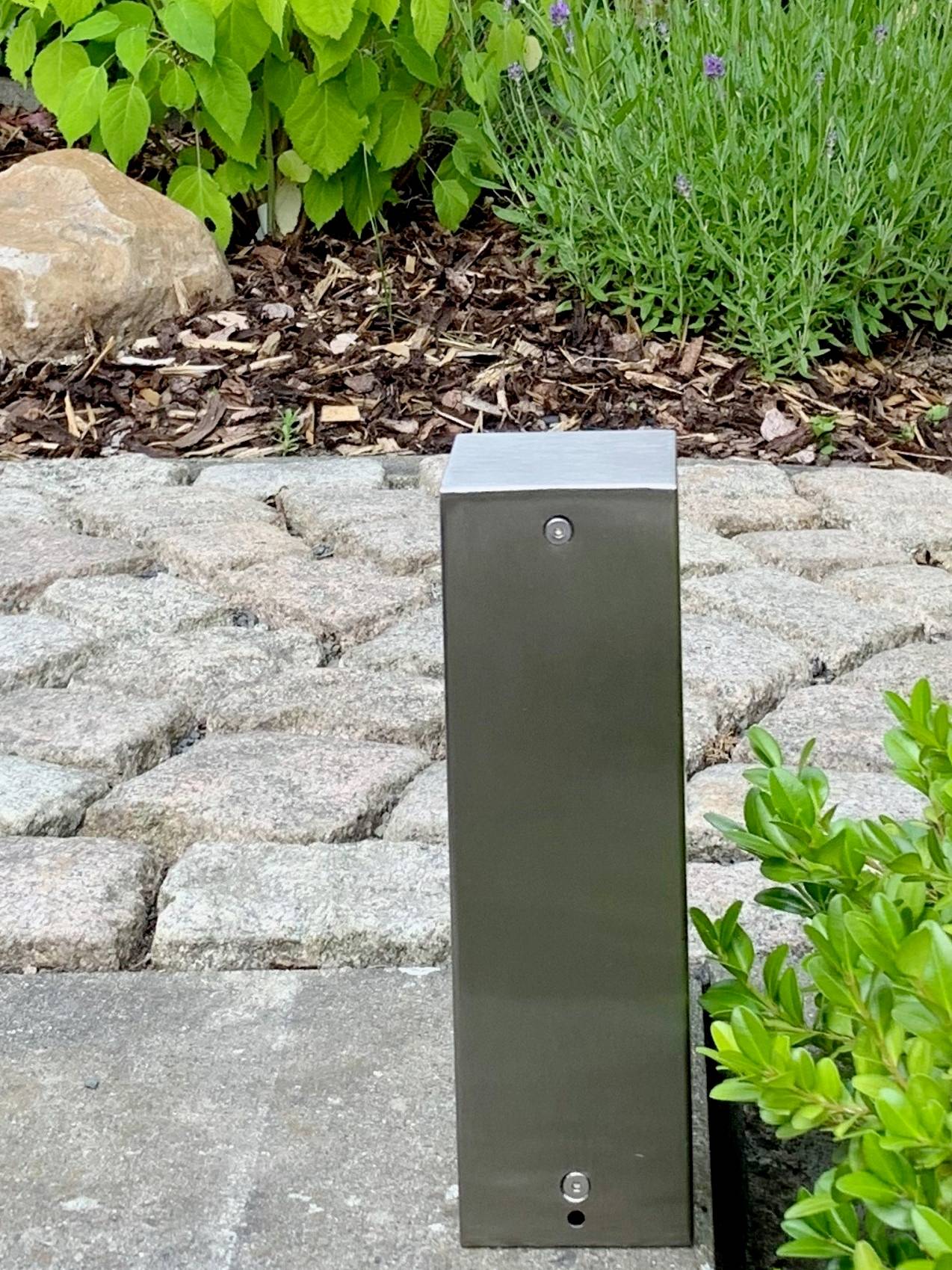 Eine niedrige, rechteckige Metallstruktur im Freien; umgeben von Pflastersteinen, Pflanzen und Erde. Trägt vermutlich zur Gartenbeleuchtung bei.