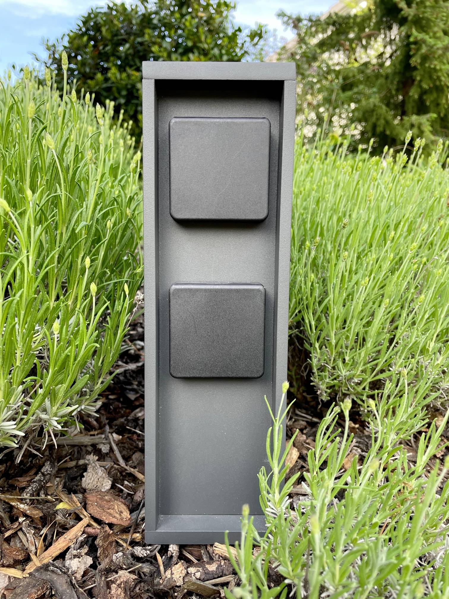 Eine graue, rechteckige Steckdosensäule mit zwei abgedeckten Steckdosen steht zwischen grünen Pflanzen in einem Garten.