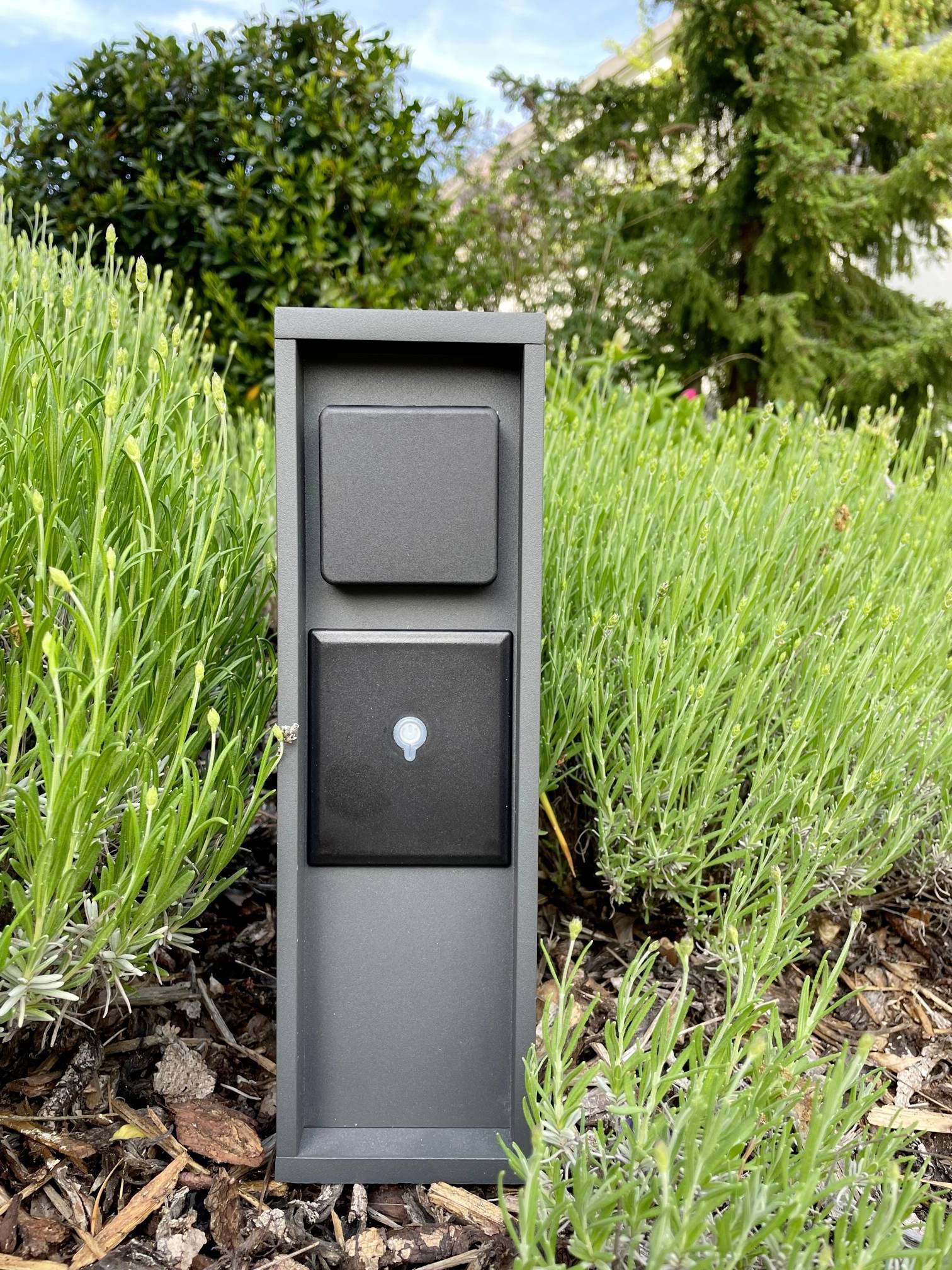 Schwarze Außensteckdose mit zwei Abdeckungen steht in einem Garten zwischen grünen Pflanzen.