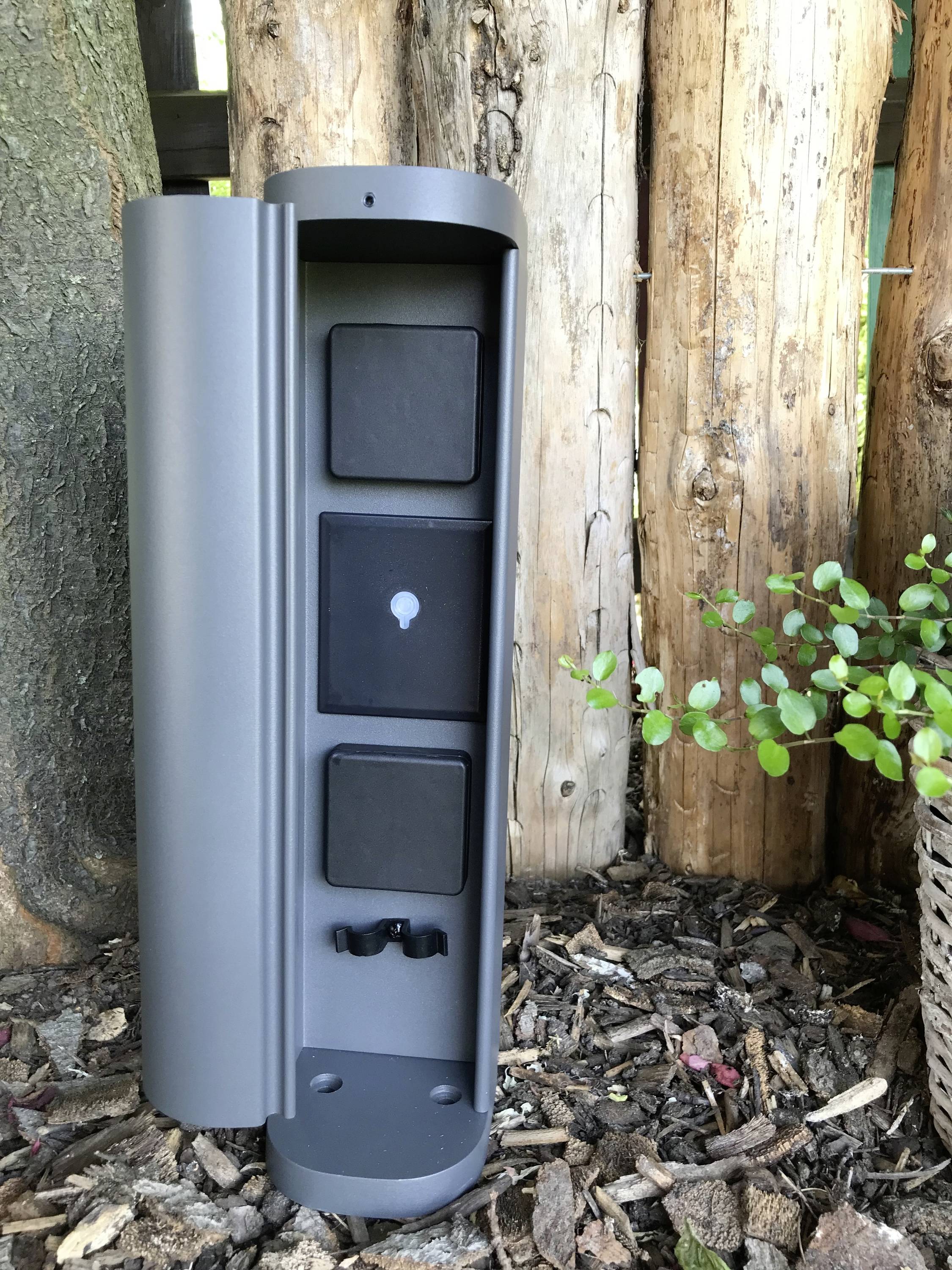 Graue Outdoor-Steckdosenleiste montiert auf Holz, umgeben von Rinde und Blättern, für Garten oder Terrasse geeignet.