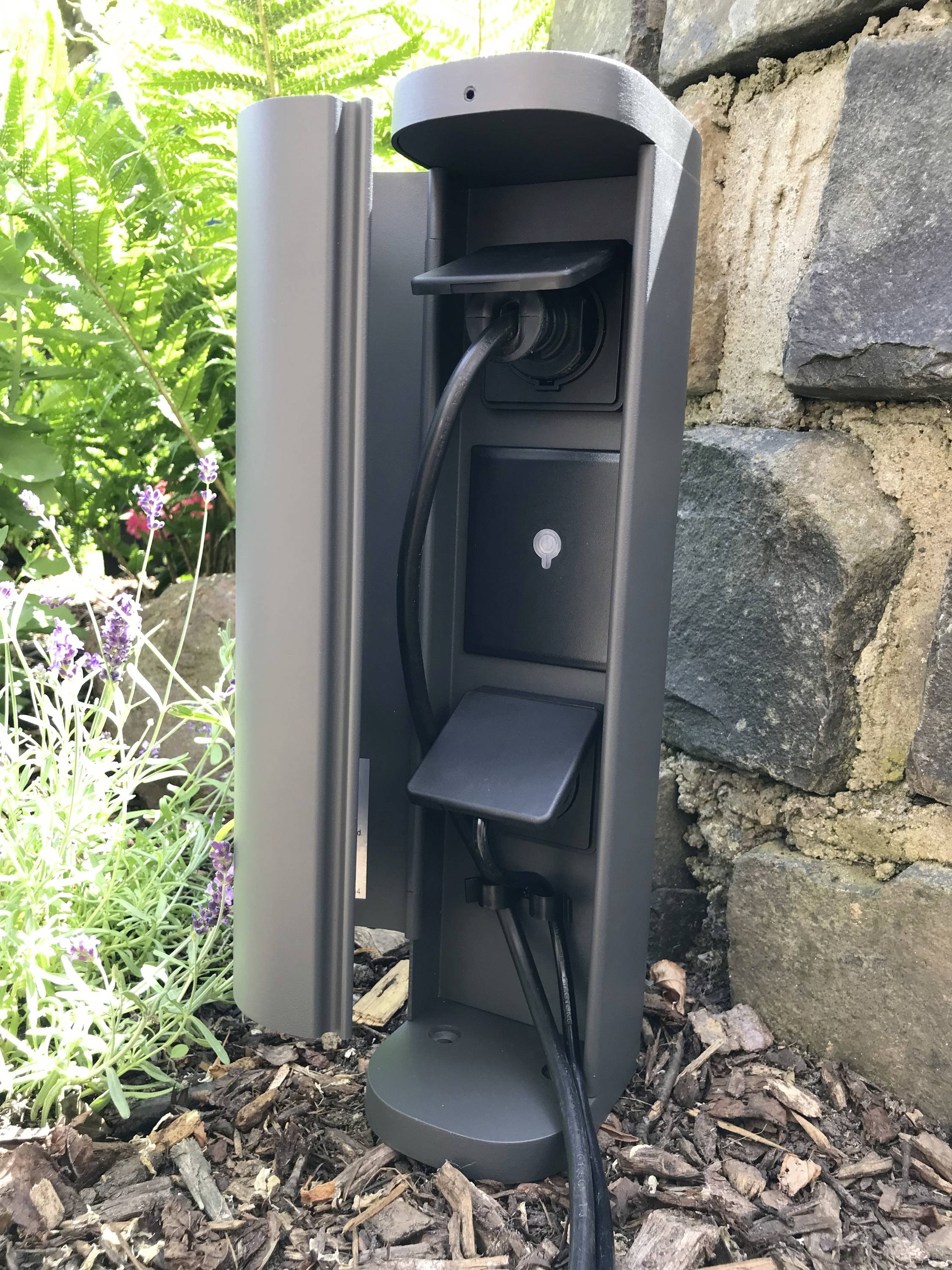 Eine graue Außensteckdose mit offenen Klappen, die in einem Garten an einer Steinmauer montiert ist, umgeben von Pflanzen.