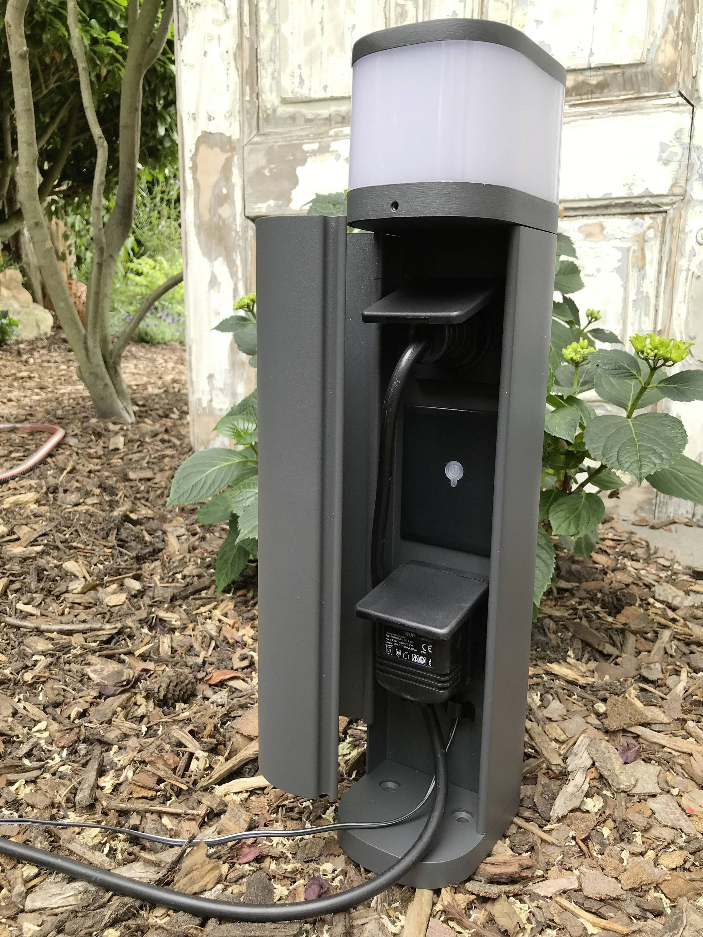 Ein grauer Außensteckdosenturm mit integriertem Licht; er steht auf Mulch mit Pflanzen im Hintergrund. Ideal für Gartenbeleuchtung.