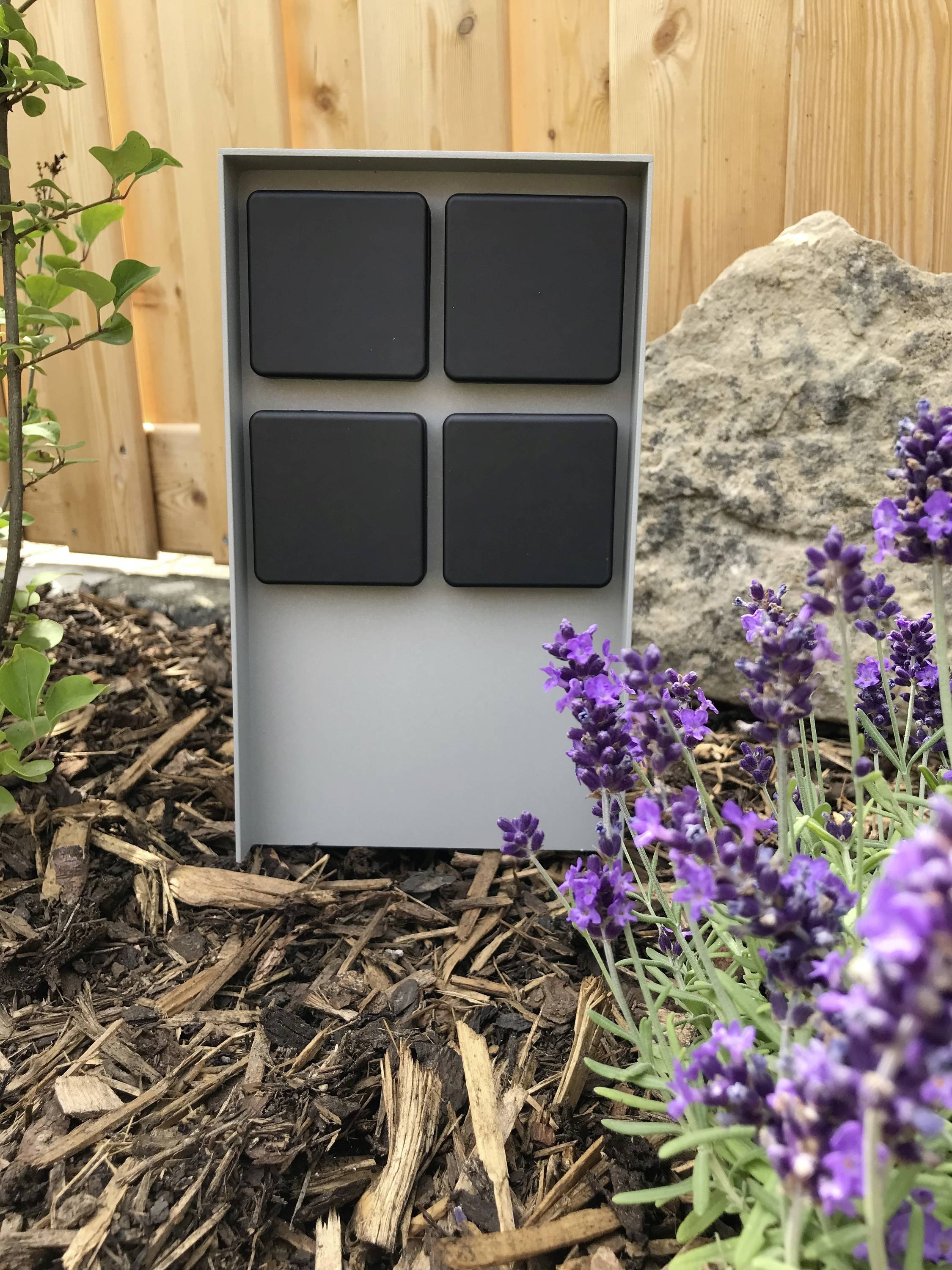 Ein graues, rechteckiges Gerät mit vier großen schwarzen Tasten steht in einem Garten, umgeben von Lavendel und einem großen Stein.