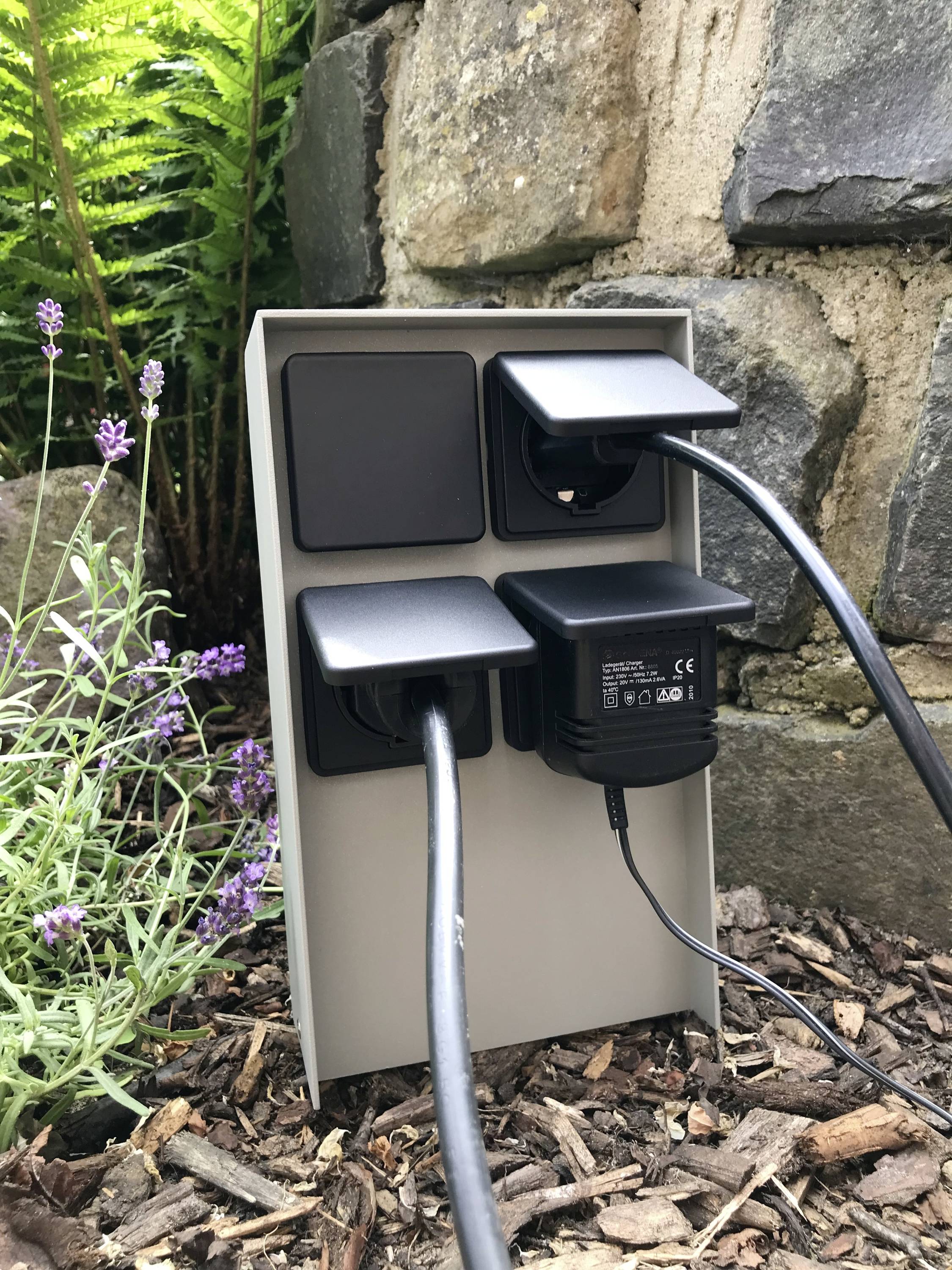 Elektrische Außensteckdose mit vier Anschlüssen, zwei davon belegt. Im Hintergrund sind Pflanzen und eine Steinmauer zu sehen.