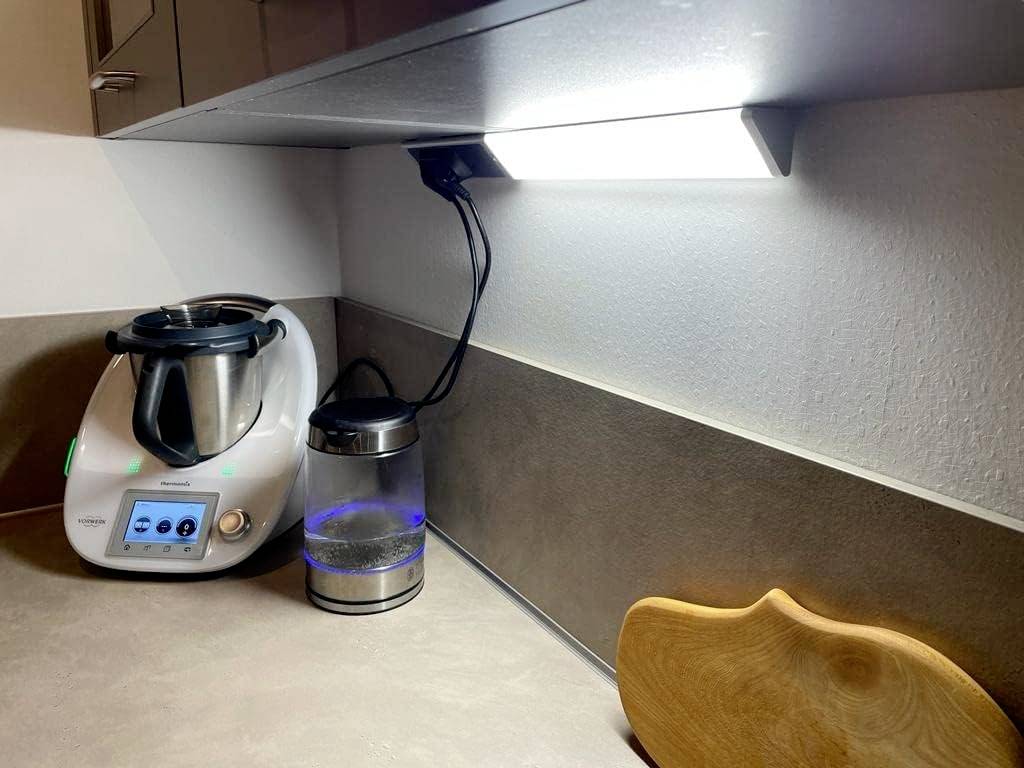 Eine Küchenzeile mit einer Kochmaschine und einem Wasserkocher unter einem eingeschalteten Licht. Daneben ein Schneidebrett.