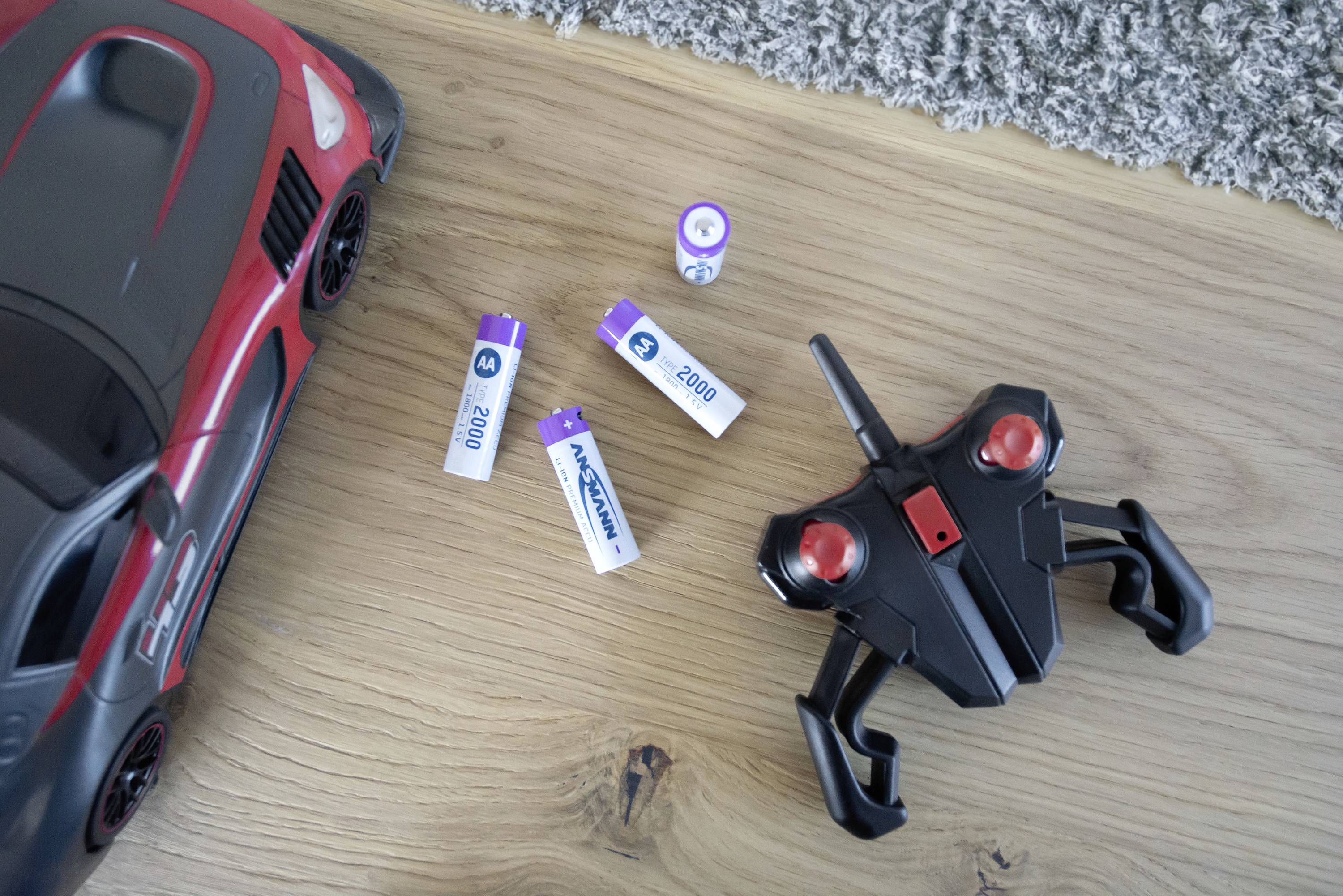 Spielzeugauto und Fernbedienung neben vier sichtbaren Batterien auf einem Holzboden. Batterien sind gekennzeichnet mit 'AA' und '2000 mAh'.